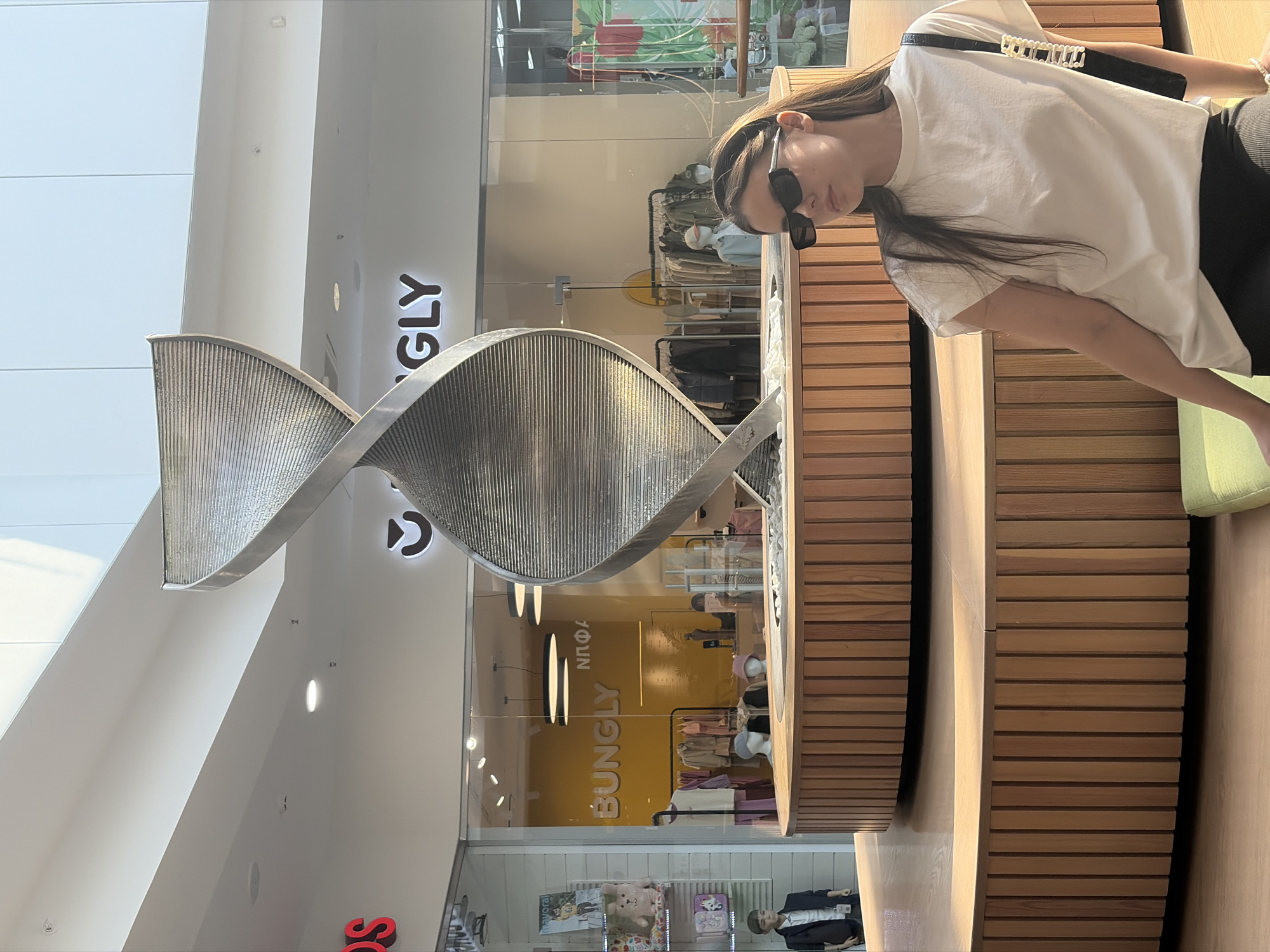 A young woman with dark sunglasses and wearing a white t-shirt stands next to a wooden slatted structure inside a shopping mall. Behind the structure is a retail space labeled "BUNGLY," and above, a distinctive twisted metal sculpture hangs under a white ceiling with skylights. The scene is brightly lit, and the architectural details, combined with the retail branding, suggest this is a modern, possibly upscale shopping environment. The location is Izhevsk, Russia.