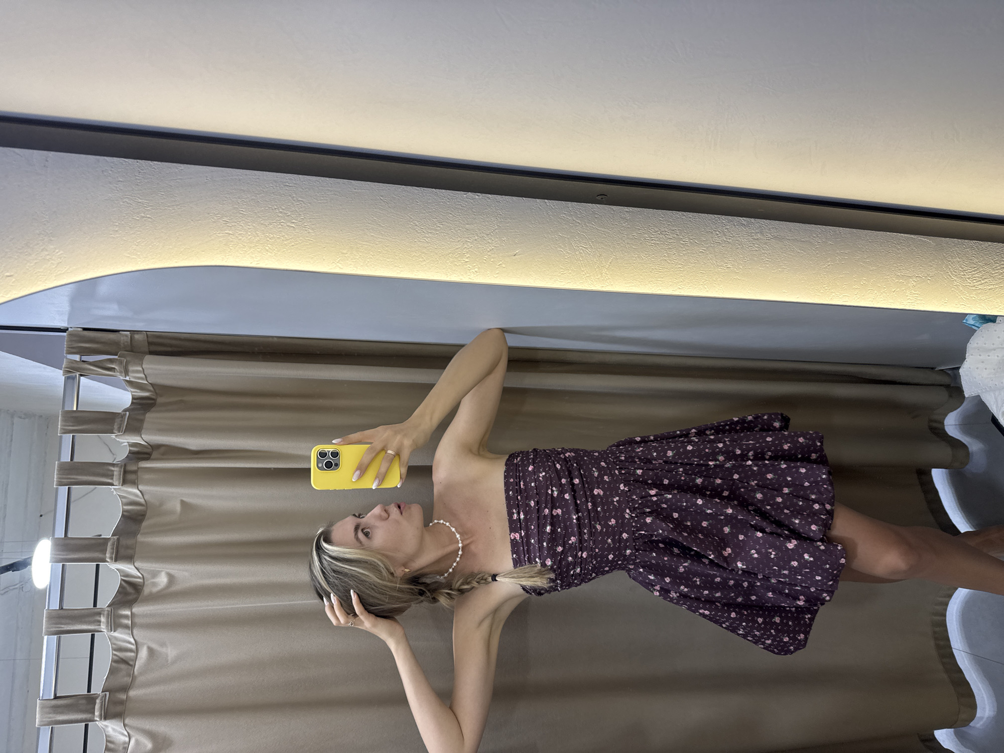 A Caucasian woman is posing for a selfie in a clothing store fitting room. She is wearing a strapless, dark purple floral print dress and a pearl necklace. Her blonde hair is styled in a braid. She is holding a yellow smartphone in her right hand and touching her hair with her left hand. The fitting room has beige curtains and a tall, illuminated mirror.