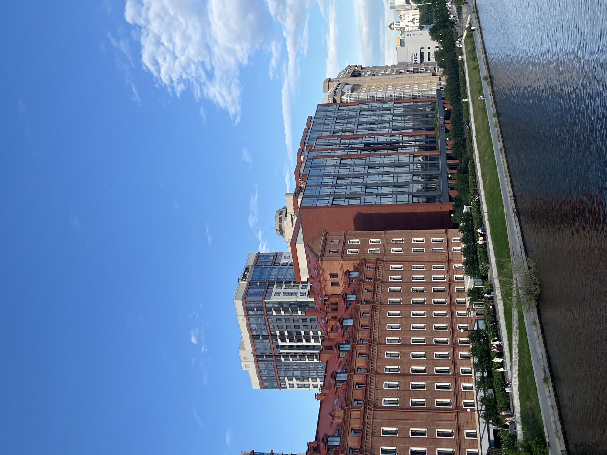 In a city scene, a body of water reflects the sky and buildings. On the right side of the frame, a modern glass-fronted building stands tall, with a brick building adjacent to it. To the left, a red brick building with a red roof stretches horizontally. Behind these structures, more buildings of varying heights and architectural styles punctuate the skyline. The sky is blue with scattered white clouds. Along the waterfront, a walkway is lined with trees and small bushes. Several people are visible walking and standing on the walkway. The overall scene depicts a mix of historical and contemporary architecture along a river.