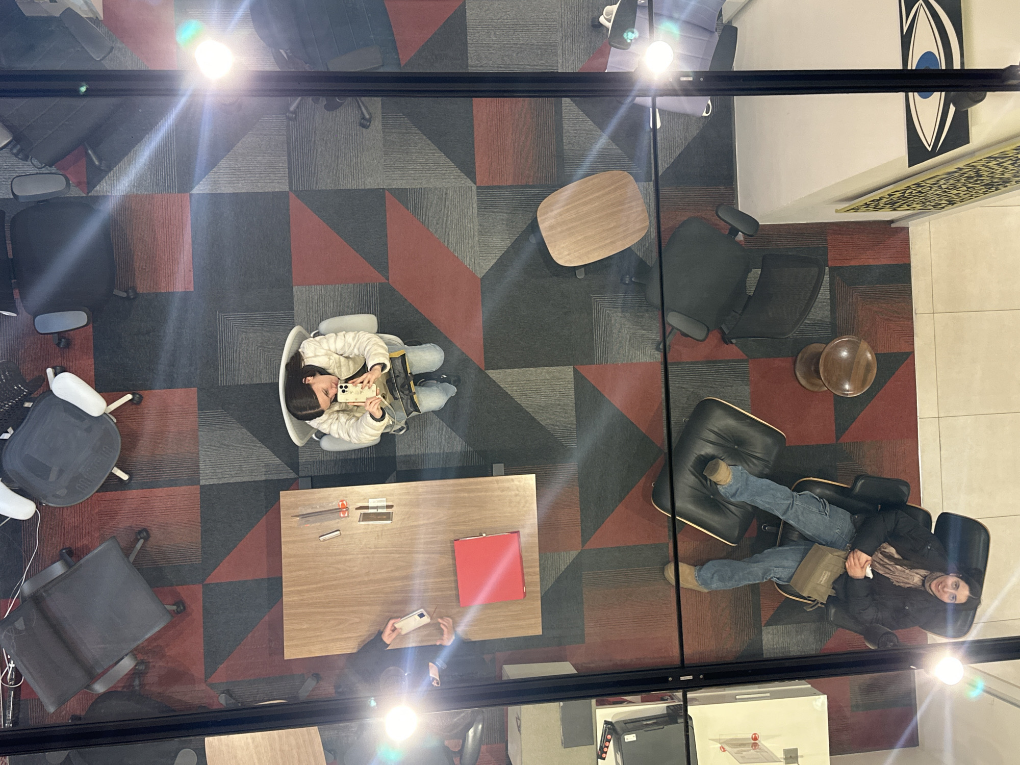 This is a high-angle view taken through a glass surface looking down into an office space. The scene appears to be in Mexico City, Mexico due to the style of art on the wall. The office has a geometric carpet with gray, black, and red patterns. There are several office chairs, including task chairs and a lounge chair, arranged around tables. Two people are present in the image, both appearing to use their cell phones. One individual is seated in a white chair, holding a phone up as if taking a photo or video. The other person is lounging in a black leather chair wearing jeans and a jacket and is looking up at the camera. On a wooden table are a red folder and other small office items. There is a bright reflection from overhead lights on the glass, causing glare in the image. The visible environment includes the office interior, with walls adorned with modern art, and office equipment such as printers visible through the glass.