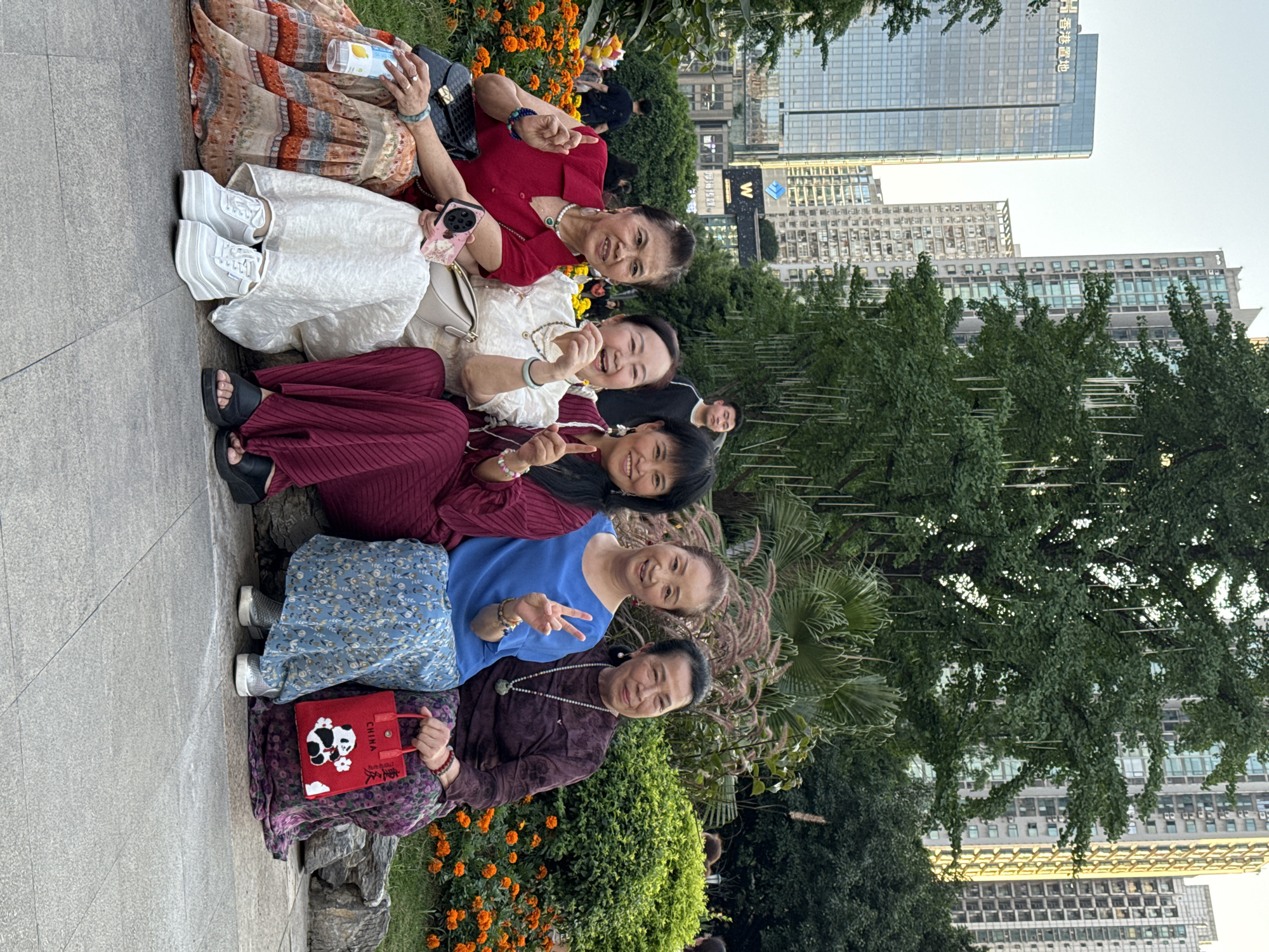 Six women are sitting side by side, posing for a photograph in Liangjiang, China. They appear to be in a park, as there are trees and flowers behind them. Several high-rise buildings of the city are in the background. The women are of varying ages and dressed in different colored clothing. The woman on the far right is holding a red bag with a panda on it. The woman next to her is making a "peace" sign with her fingers. The woman at the top is holding a mobile phone and a bottle.