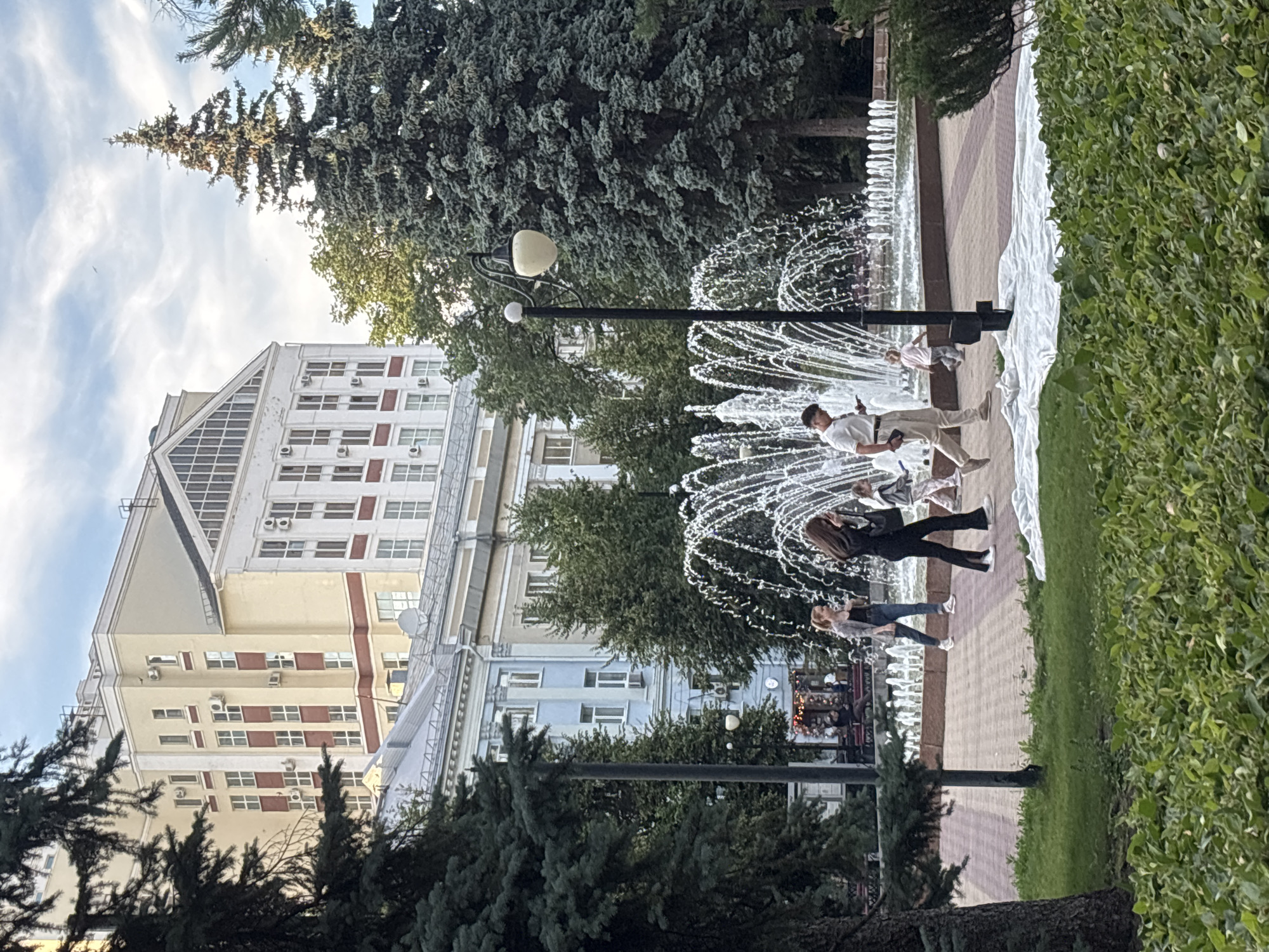 A daytime, eye-level, outdoor shot captures a public park area with a fountain and several people walking by. In the background, a multi-story building with a cream-colored facade, red accents, and numerous windows stands prominently. Adjacent to it, a lighter colored building with a large triangular architectural element is visible. To the left, a light blue building with windows and decorative architectural details is present, partially obscured by trees. In the middle ground, a large, active fountain sprays water upwards, creating a misty effect. People are walking on a patterned walkway in front of the fountain. One individual in a white polo shirt and beige pants is looking at their phone, while another person in a black outfit and a third in a grey jacket are also walking. A young child is visible near the fountain. The foreground features lush green grass and dense shrubbery. Tall evergreen trees frame the scene on the left and right. The sky above is partly cloudy with a pale blue hue. The location is identified as Novaya Usman', Russia, specifically within a park area likely adjacent to urban architecture.