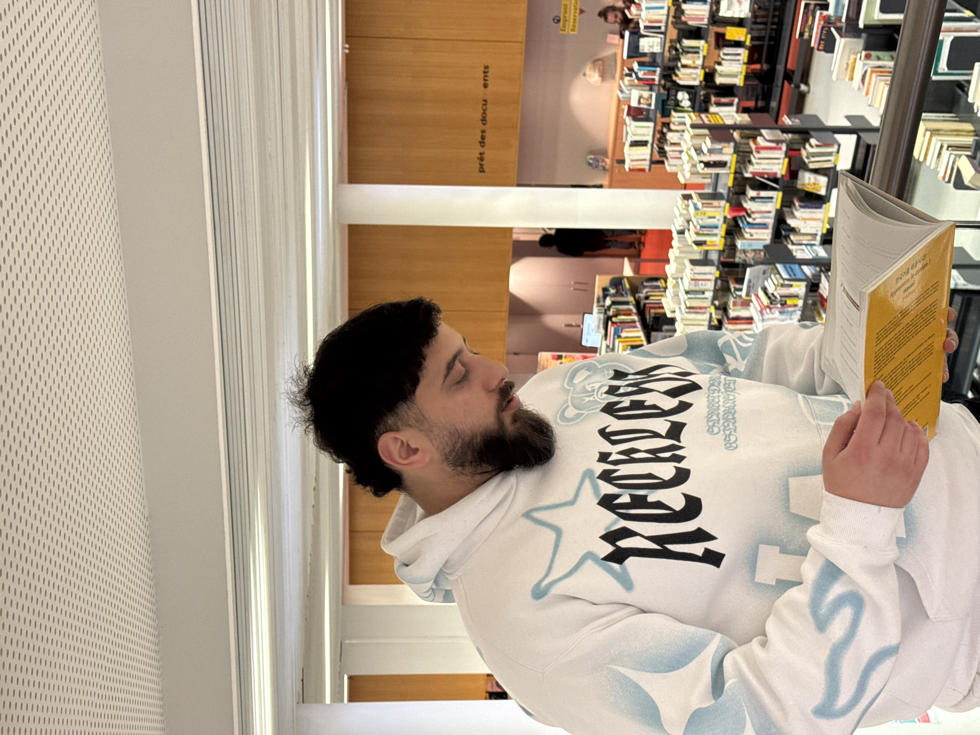 A man is reading a book in the Bibliothèque nationale et universitaire de Strasbourg, a large public library located in Strasbourg, France. The man has dark hair and a beard, and he is wearing a white hoodie with black lettering and blue designs. He is holding the book in his hands and looking at the pages. Behind him, there are bookshelves filled with books. In the background, there is a sign that says "prêt des documents".