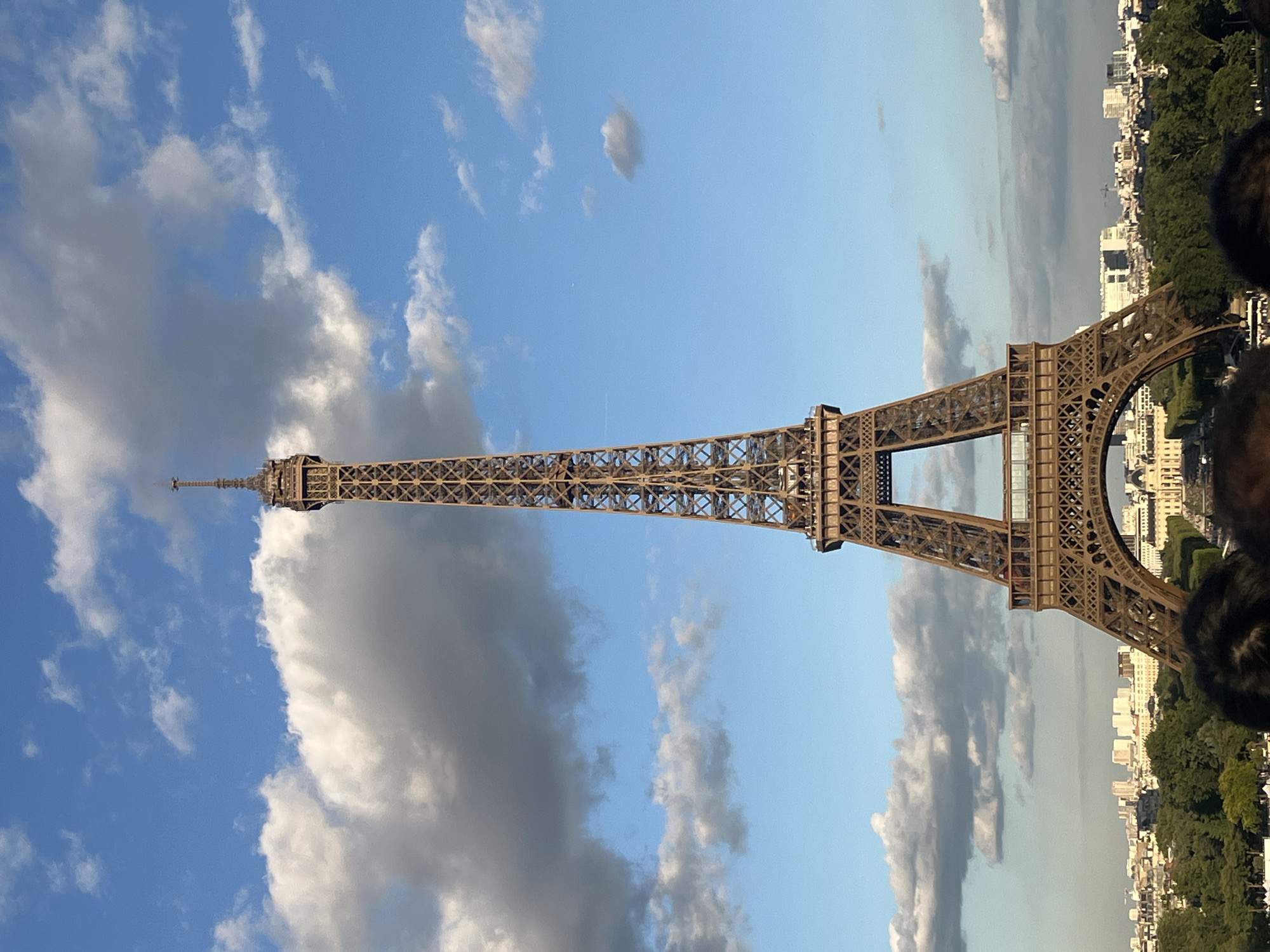 This is a full-frame, eye-level, medium shot photograph of the Eiffel Tower in Paris, France, taken on a clear day with a partially cloudy sky. The Eiffel Tower dominates the center of the frame, its intricate iron latticework a prominent feature against the bright blue sky dotted with white and grey clouds. The tower is a warm, reddish-brown color, and its base is partially obscured by lush green trees and the tops of people's heads in the foreground, indicating the photo was taken from a raised vantage point or a crowd. Beyond the tower, a cityscape of Paris stretches across the horizon, characterized by low-rise buildings and some taller structures in the distance. The lighting suggests it is daytime, possibly late afternoon, with the sun casting a warm glow.