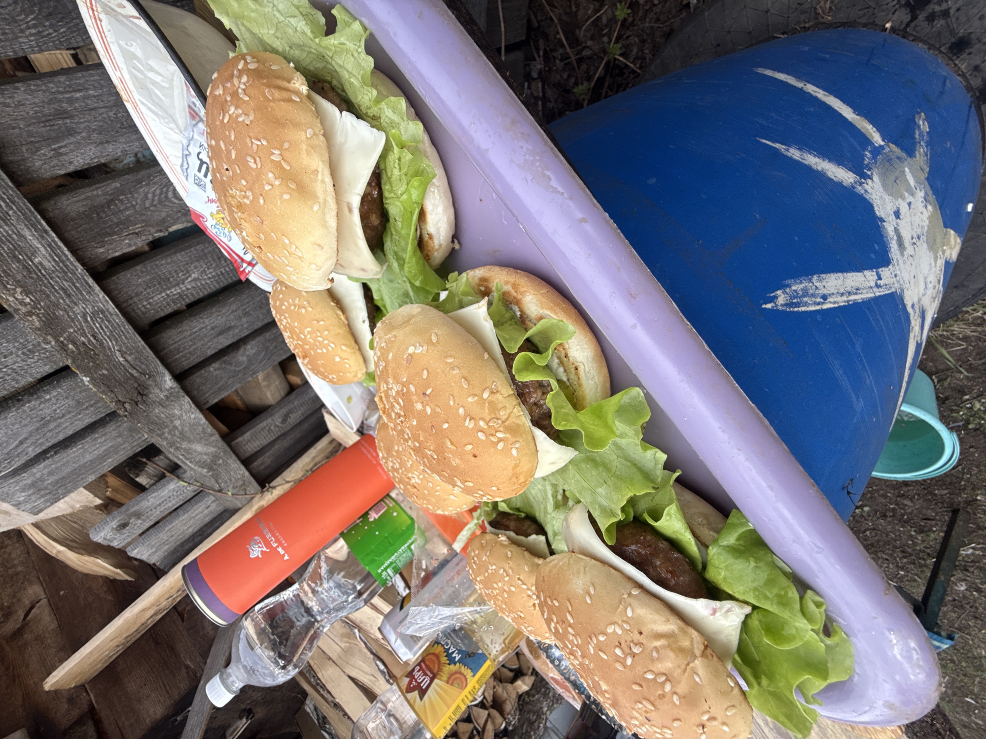 The image displays an outdoor scene, likely a casual meal in Кемуль, Russia. Six or more elongated burgers or hot dogs, featuring sesame seed buns, lettuce, and white cheese slices, are presented within a long, light purple plastic container. This container is resting on a surface composed of weathered wooden planks.
To the left of the container, a white paper bag with red and yellow designs is partially visible. An orange cylindrical object, possibly a thermos or insulated bottle, stands nearby. Visible text on this object includes "A. DE FUSE" and "COLLECTION," along with some indistinct Cyrillic script. Beside it is a clear plastic bottle, likely containing water. Below these items, a yellow box with a prominent sunflower image and Cyrillic text reading "МАСЛО ЦАРЬ ПОДСОЛНЕЧНОЕ" (Sunflower Oil "Tsar") is present among the wooden planks.
In the background, a large blue barrel with several white, streaky paint marks is visible. A small, light green plastic bucket can be seen near the base of the blue barrel. The ground beneath appears to be dirt with sparse green vegetation. The scene suggests an informal outdoor gathering or meal preparation during daylight hours.