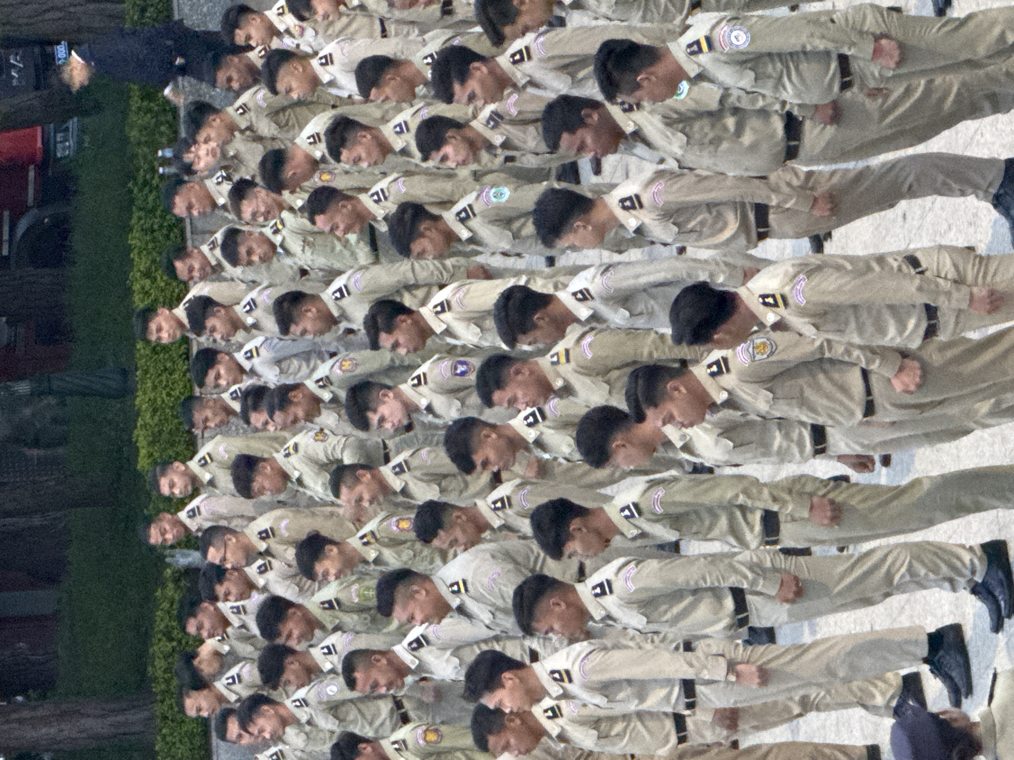 A group of young men in khaki uniforms stand in formation outdoors. The uniforms consist of long-sleeved shirts with epaulets and patches on the shoulders, and khaki pants with black belts. Most of the men have short haircuts. The men are standing on a paved surface, and a low hedge and trees are visible in the background. There is a red vehicle in the upper right corner of the image and a person in a dark suit walking in the background. The scene appears to be a formal gathering or ceremony. While the image does not explicitly state the location is Da Lat, Vietnam, the presence of individuals in uniforms and the overall setting could be consistent with a location in Vietnam.