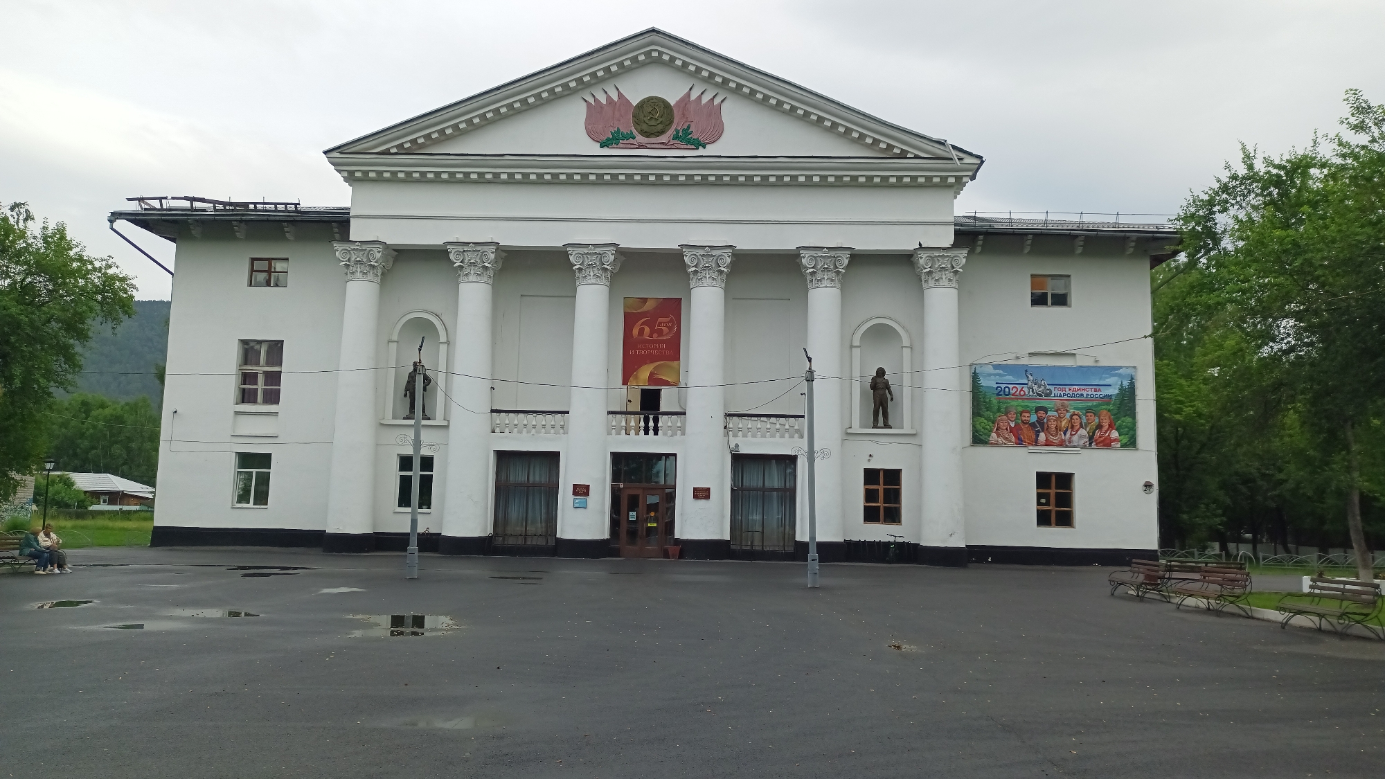 The image depicts the Abaza City Palace of Culture, a white, two-story building with classical architectural elements, including a pediment topped with a Soviet-era emblem and several large columns supporting a portico. A banner celebrating "65 years of creativity" hangs between two columns, and a mural depicting people in traditional attire with the text "2026 GOD EDINSTVA NARODOV ROSSII" (2026 Year of Unity of the Peoples of Russia) is displayed on the right side of the building. A statue of a person stands in an alcove on the upper floor. In front of the building, a paved area with puddles suggests recent rain. Two individuals are seated on a park bench on the left, facing away from the building. To the left of the building, a small residential house and trees are visible, and to the right, more trees line the perimeter. The sky is overcast.