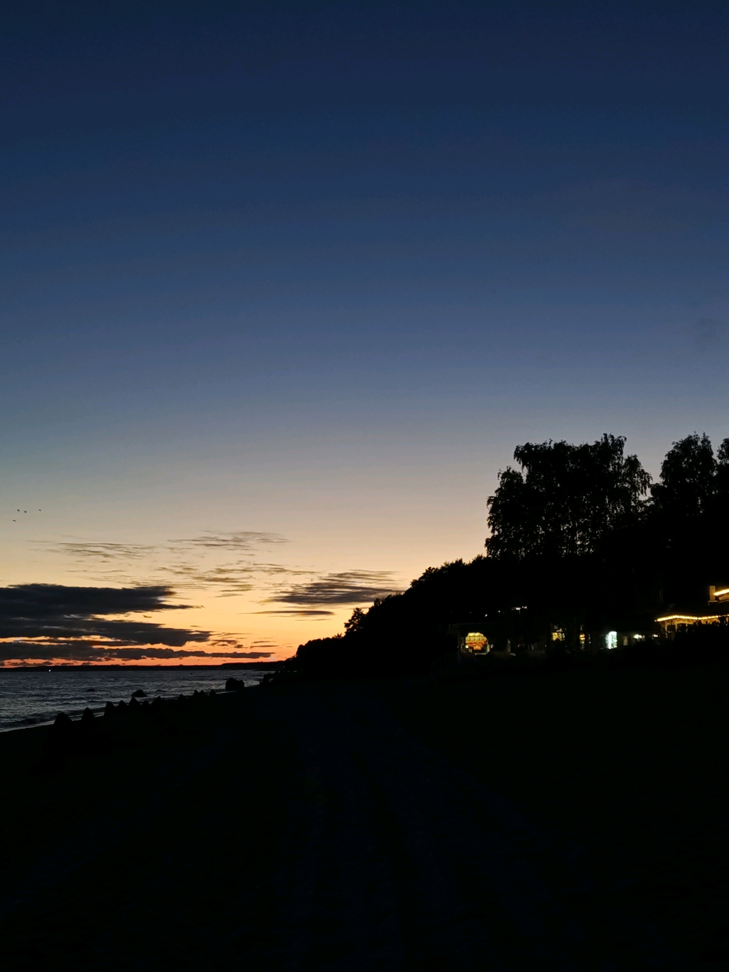 The image depicts a twilight scene at the Komarovo beach in Komarovo, Russia. The sky transitions from a deep blue at the top to hues of orange and yellow near the horizon, indicating either sunrise or sunset. Several dark clouds are visible, adding texture to the sky. The sea is dark and appears calm, with gentle waves lapping at the shore.
The foreground is dominated by the silhouetted shape of the beach and a dark, tree-covered incline. On the incline, there are faint lights from what appears to be buildings or structures, possibly a cafe or restaurant, suggesting human activity. A few small birds are flying in the distance, near the horizon. The overall atmosphere is serene and quiet, characteristic of a beach at dusk.