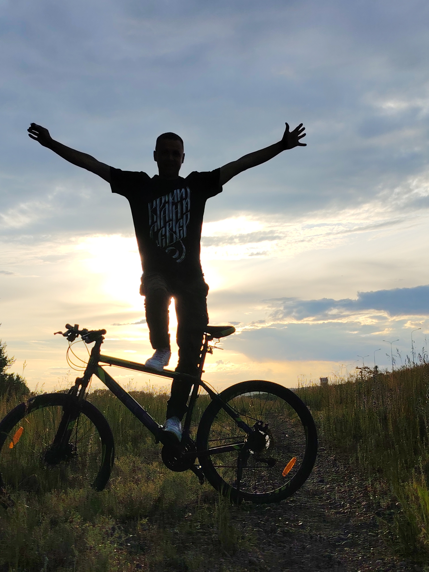 A person is standing on a bicycle with their arms outstretched against a cloudy sky at sunset. The sun is visible behind the person, casting a silhouette. The person is wearing a dark t-shirt with white lettering and dark pants, and white sneakers. The bicycle is dark-colored with a green accent. The scene is set in a grassy, outdoor environment with a dirt path. In the background, there are trees and utility poles. The time of day is sunset, indicated by the low sun and the colors in the sky. The weather appears to be partly cloudy. The image was taken in Chaikovsky, Russia. The person's pose and outstretched arms suggest a feeling of freedom or triumph. The text on the t-shirt is difficult to read due to the silhouette, but appears to be in a stylized font.