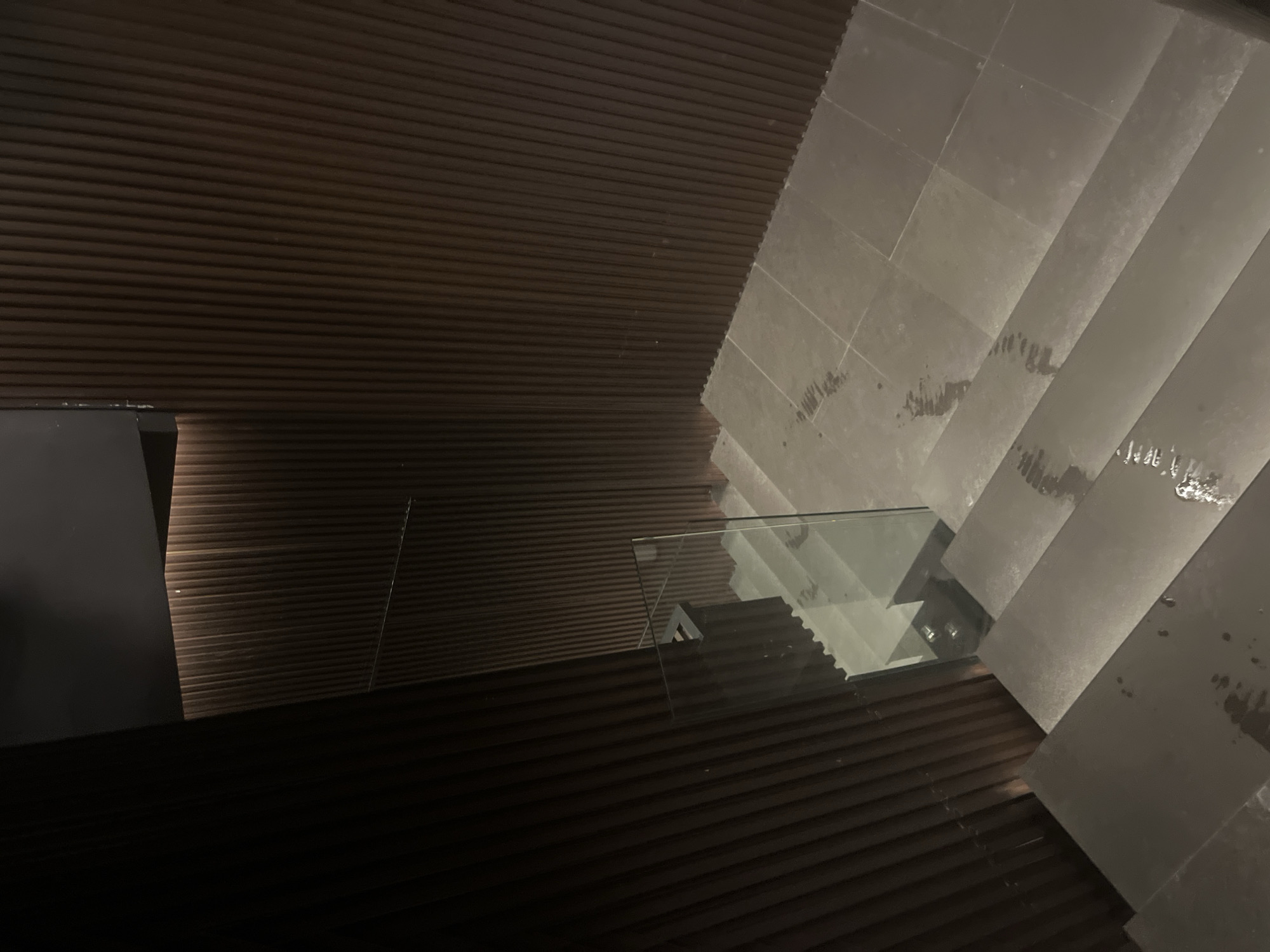 This image presents a modern architectural interior, likely a building in Sofia, Bulgaria, captured from an elevated perspective looking downwards. The scene is characterized by minimalist design and contrasting textures.
The left and bottom portions of the frame are dominated by a dark brown surface featuring numerous thin, horizontal slats or grooves, creating a textured, linear pattern that suggests a wall or paneling. Towards the far left, a dark, unidentifiable architectural element or structure is partially visible.
Diagonally across the middle of the image, a transparent glass railing or balustrade acts as a visual separator. The glass reflects parts of the surrounding environment, including the stairs and the slatted wall, adding depth and complexity to the composition.
On the right side, a wide, modern staircase descends. The steps are made of a light grey material, possibly concrete or stone, and have a clean, block-like appearance. Some darker, irregular marks or smudges are visible on the surface of the steps.
The lighting in the scene is dim and artificial, creating a subdued atmosphere and highlighting the material textures. No people are visible, and there is no discernible activity taking place. The overall impression is one of contemporary design, quietude, and a focus on architectural form and material. No text is visible within the image.