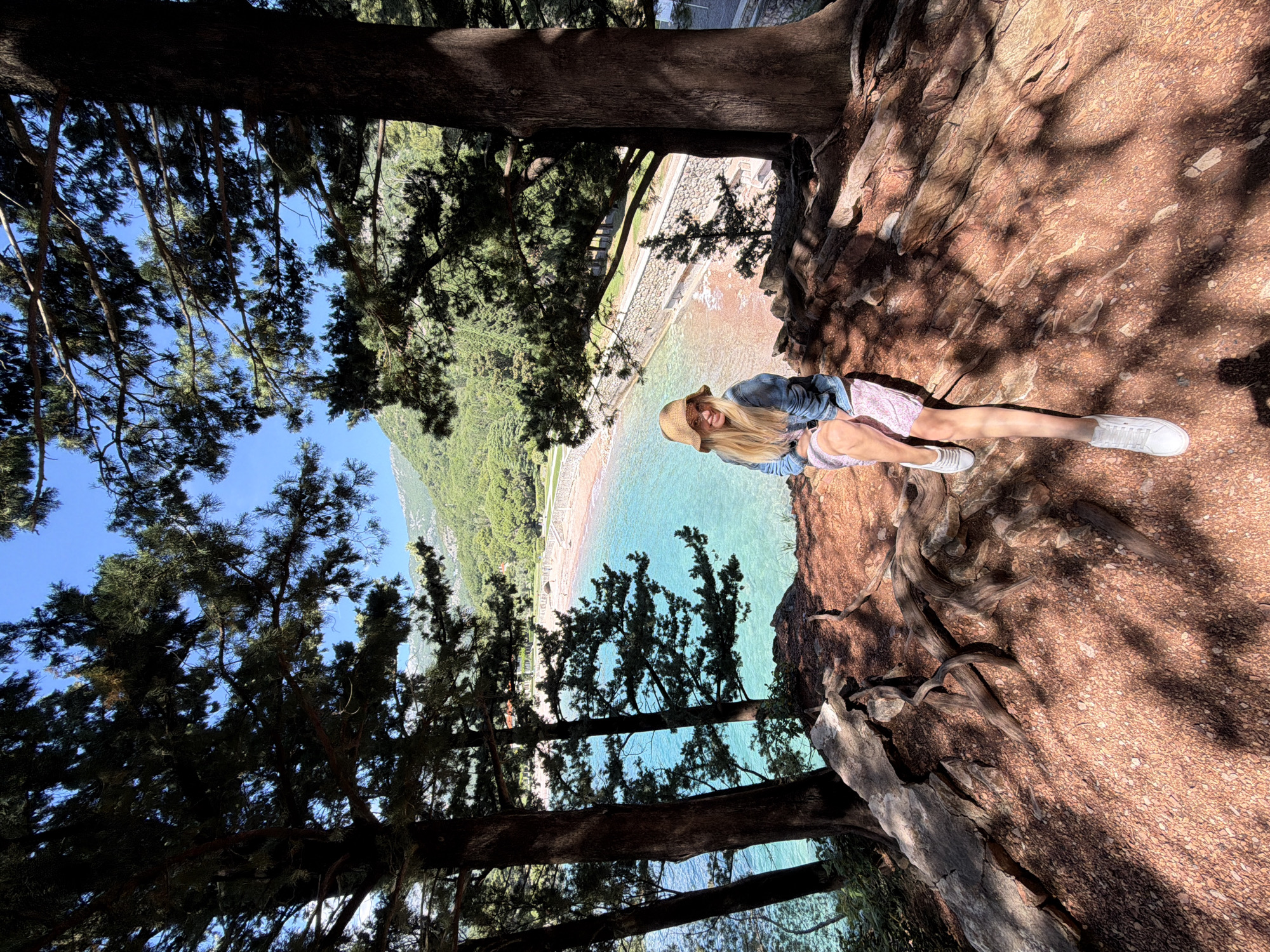 A panoramic shot taken from a high vantage point in Sveti Stefan, Montenegro, features a woman lying on a reddish-brown, rocky surface. She is wearing a straw hat, a denim jacket, and a patterned skirt. Her legs are extended, and she wears white sneakers. The background reveals a coastline with turquoise water, a sandy beach, and lush green vegetation. The shot is framed by trees, with branches and leaves occupying the upper and lower portions of the image, casting shadows and creating a natural enclosure around the central scene.