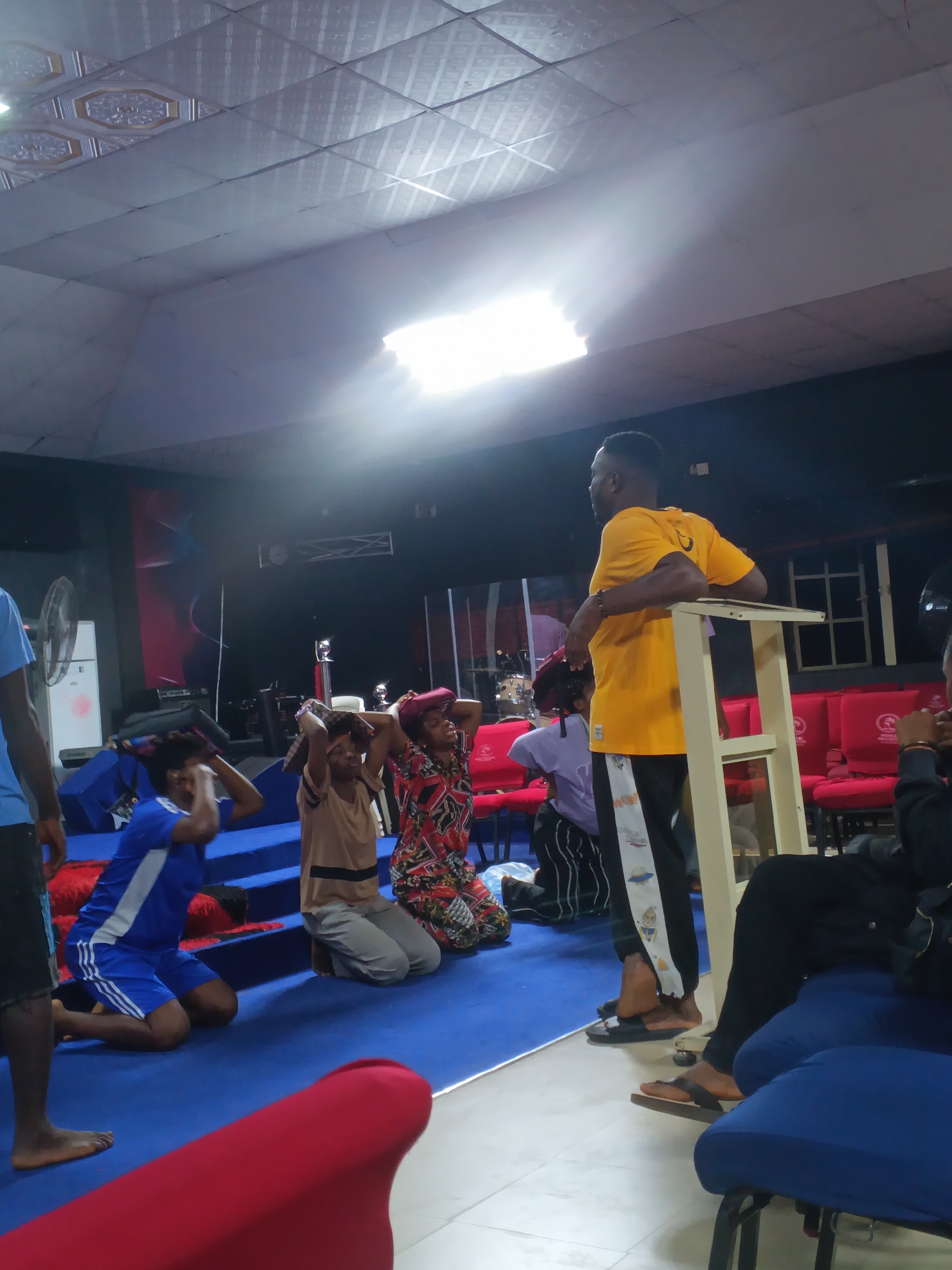 In a room with a suspended white tiled ceiling, several people are gathered on a blue carpeted floor. A man in a bright yellow t-shirt stands on the right, leaning on a white structure. In the center foreground, a man in a blue and white athletic outfit kneels with his hands on his head, alongside a woman in a tan top and grey pants also kneeling with her hands on her head. To the right of them, another person in a colorful patterned shirt and a head covering kneels with their hands on their head. A person in a purple shirt and striped pants is also kneeling in the background. The environment appears to be a hall or church, with rows of red chairs visible in the background, along with a drum set, a fan, and a whiteboard. The overall lighting suggests an indoor setting with artificial illumination. This scene is observed in Nkpor, Nigeria.