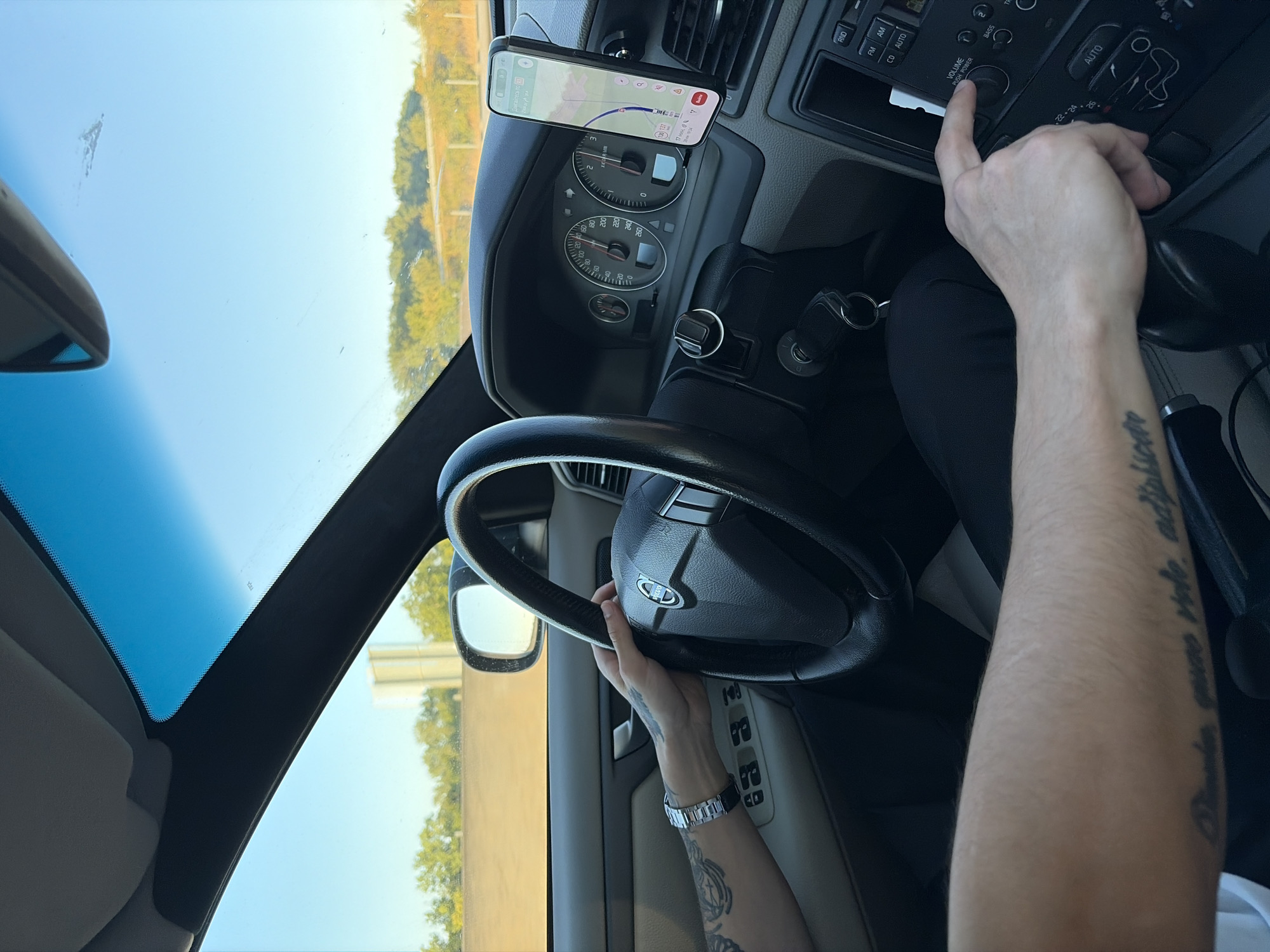 A first-person perspective shot from inside a car shows the driver's hands on the steering wheel and operating the car's audio system. The driver's left hand, adorned with tattoos and a watch, grips the steering wheel. The driver's right hand is adjusting the volume knob on the car's dashboard. The dashboard displays the car's speedometer and a phone mounted on the windshield showing navigation. The exterior of the car, visible through the windshield, indicates a clear day with trees and a highway. The scene is consistent with a person driving in Dolná Streda, Slovakia.