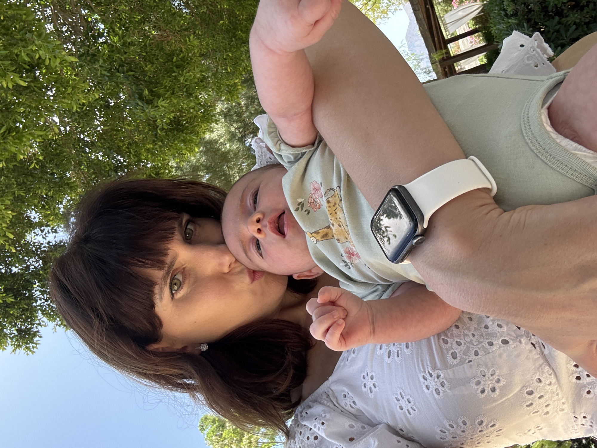 A woman with dark hair and bangs is holding a baby in her arms outdoors. The woman is kissing the baby on the forehead. The baby is wearing a light green onesie with a pattern of animals and flowers. The woman is wearing a white top with eyelet detailing. The background consists of trees, foliage, and a clear blue sky. The scene appears to be in a garden or park-like setting. The woman is wearing a smartwatch on her left wrist. The overall impression is of a tender moment between a mother and child.
