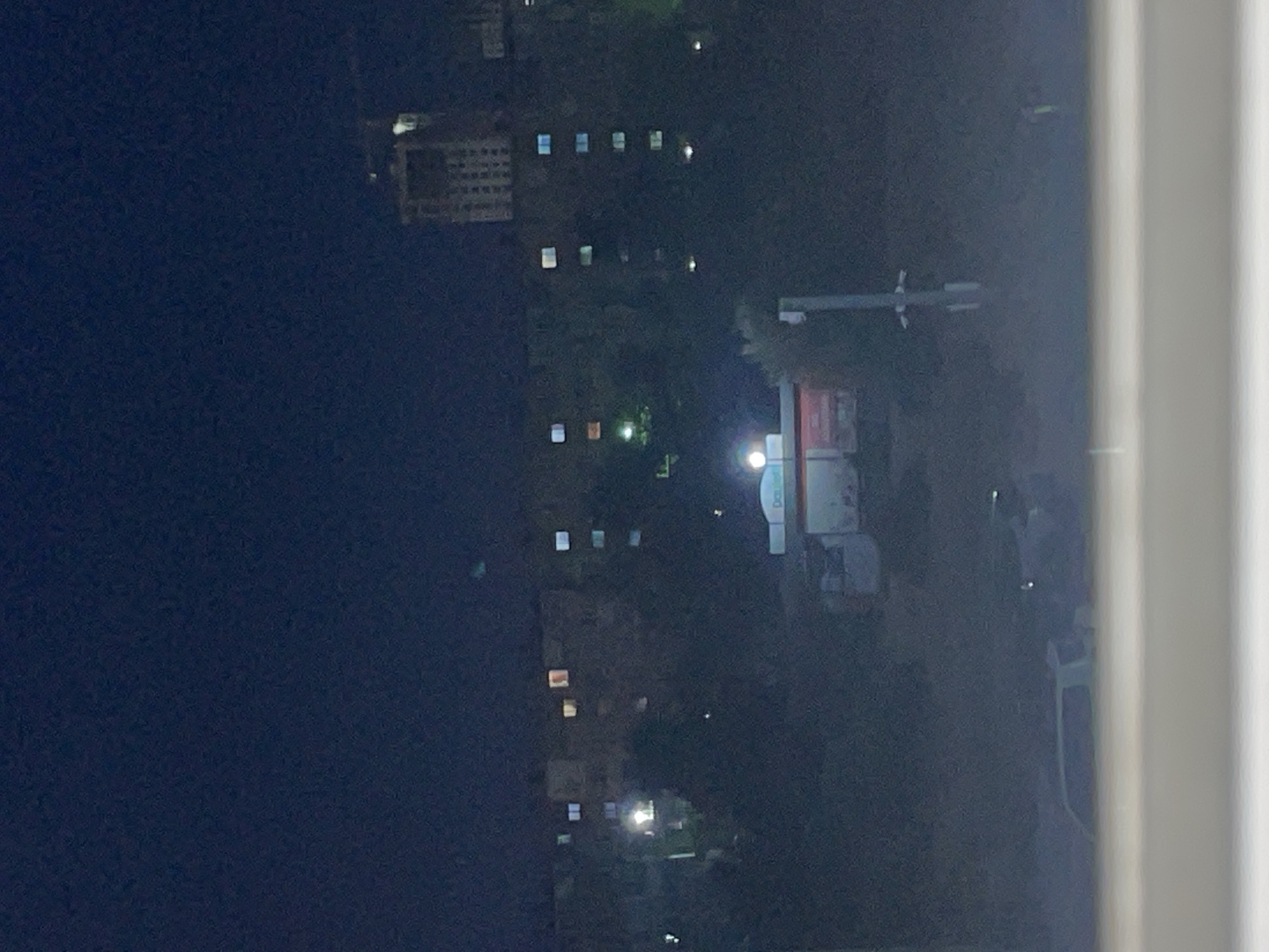 The image shows a nighttime view of a city scene in Aktau, Kazakhstan. In the background, a tall, unfinished building with a crane on top is visible. In front of this building are several apartment blocks with illuminated windows. In the midground, there is a small structure with lights on, possibly a kiosk or shop, surrounded by trees. In the foreground, the silhouette of a car is visible, along with what appears to be a road or pathway. The overall scene depicts a residential area at night.