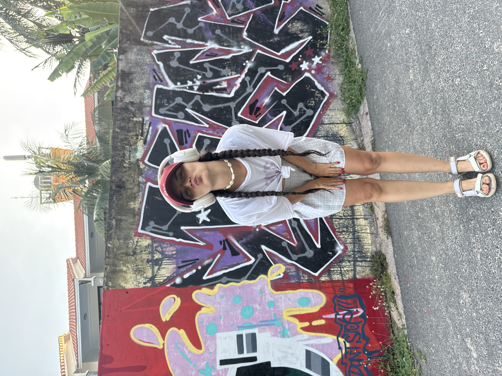 A young woman with long, braided dark hair and wearing white headphones, a white t-shirt, a red baseball cap, and light-colored checkered shorts is posing for the camera in Phu Thuy Ward, Vietnam. She is standing on a paved surface next to a graffiti-covered wall. The graffiti features various colors, including purple, black, white, and red, with abstract shapes and bold lettering. To the left of the wall, there are palm trees and a building with a red tiled roof. The woman is wearing white sandals with light straps. Her expression is playful as she puckers her lips towards the camera. The lighting suggests it is daytime, possibly late afternoon, with overcast skies. The scene appears to be an outdoor urban environment with a touch of tropical greenery.