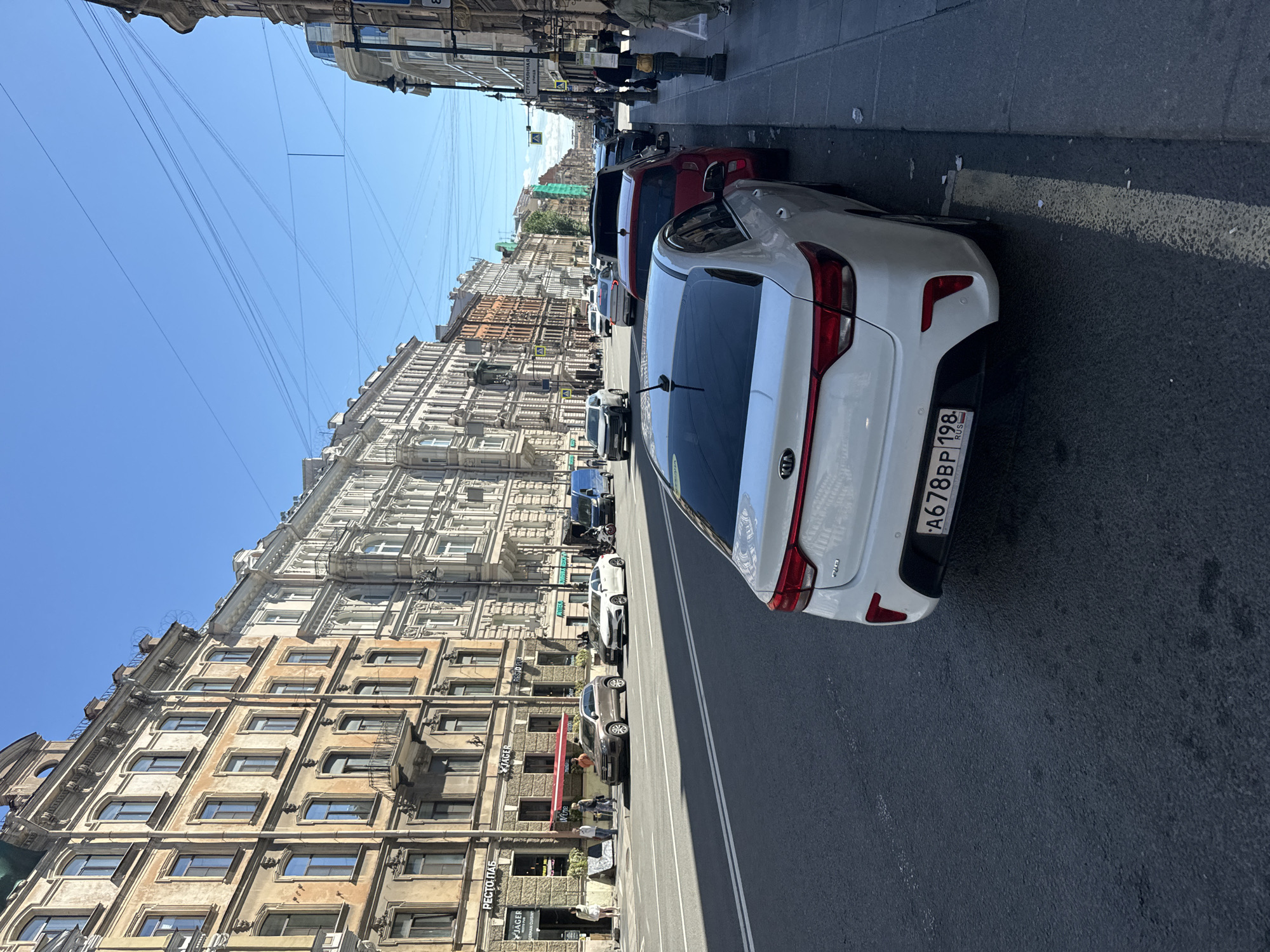 A wide street in Saint Petersburg, Russia, is lined with historic buildings and parked cars. The buildings, with their ornate facades and numerous windows, appear to be from the 19th or early 20th century. Several cars are parked along the right side of the street, and others are driving in the distance. The sky is clear and blue, indicating a sunny day. A few pedestrians can be seen on the sidewalks. The overall scene depicts a typical urban streetscape in a Russian city with a rich architectural heritage.