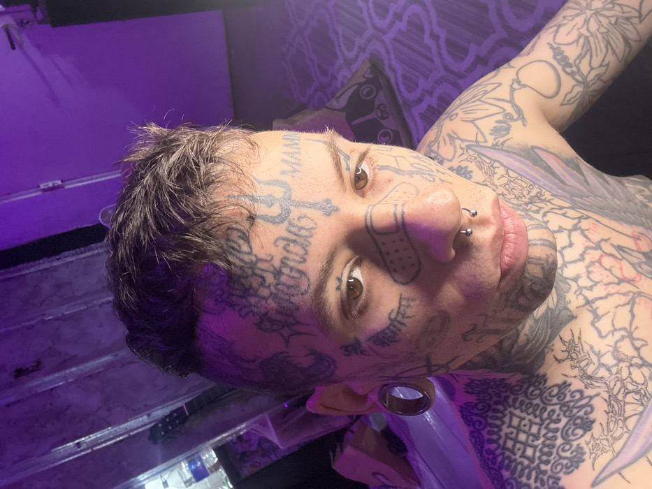 An individual is depicted in a close-up, chest-up indoor portrait, likely a selfie, under predominantly purple lighting. The exact location within Buenos Aires, Argentina, is not discernible from the image.
The subject is a young person with short, dark, textured hair and light brown eyes, looking directly forward. Their face, neck, and upper chest are extensively covered with numerous tattoos, appearing to be black or dark grey ink. Notable facial tattoos include stylized script and symbols across the forehead, temples, and cheeks. A prominent trident-like symbol is tattooed on the center of the forehead. On the bridge of the nose, a tattoo resembling a beige-colored adhesive bandage with ventilation holes is visible. The subject has a silver septum piercing with spherical ends and large, circular ear gauges in both earlobes, visible on the left side. Text tattoos like "MAMB" are present on the right temple, and symbols resembling Devanagari script are visible on the left cheek.
The upper torso displays dense, intricate tattoo patterns, including geometric designs, floral motifs, and abstract shapes, extending down the arms. The individual is shirtless, revealing the extensive body art.
The background is dimly lit and largely out of focus, dominated by a pervasive purple hue. On the left side, a clear plastic water bottle and a partial view of what appears to be a guitar neck are faintly visible. On the right, a patterned textile, possibly a pillow or blanket with a repeating circular or geometric design, is present. The overall environment suggests a private, personal space.
