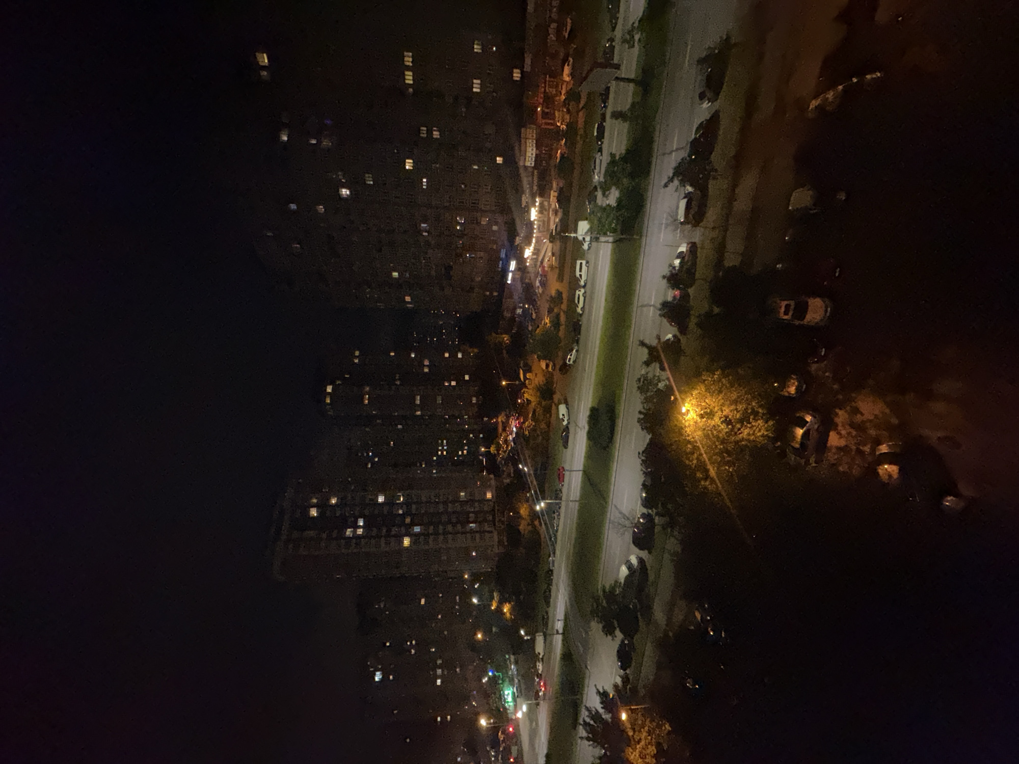 The image captures an elevated nighttime view of an urban environment in Kyiv, Ukraine, likely from an apartment building. In the foreground, several cars are parked on a darkened, possibly tree-lined street. The middle ground shows a multi-lane road with sparse vehicular traffic, indicated by headlights and taillights, and streetlights illuminating the asphalt. On the far side of the road, more parked cars and some greenery are visible. Dominating the background are large, multi-story apartment buildings, their windows illuminated by interior lights, creating a grid-like pattern against the dark night sky. The overall atmosphere is one of a quiet urban evening.