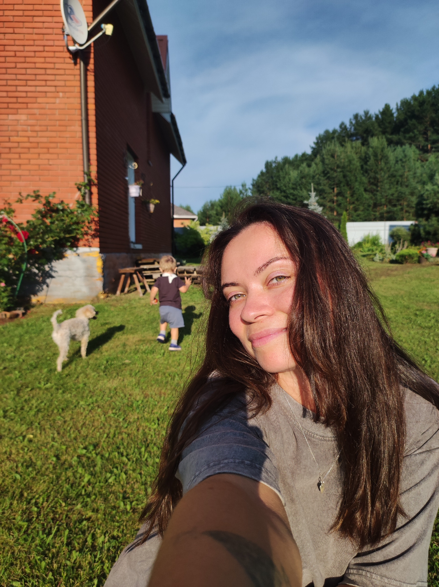 A woman with long, dark hair takes a selfie outdoors in a grassy yard. She is wearing a gray t-shirt and a silver necklace. In the background, a young child in a dark shirt and blue shorts walks away from the camera, and a small, light-colored dog stands in the grass. A brick house with a satellite dish is visible to the left, and a line of trees forms the background. The scene appears to be in the city of Fokino, Russia, given the metadata associated with the image.