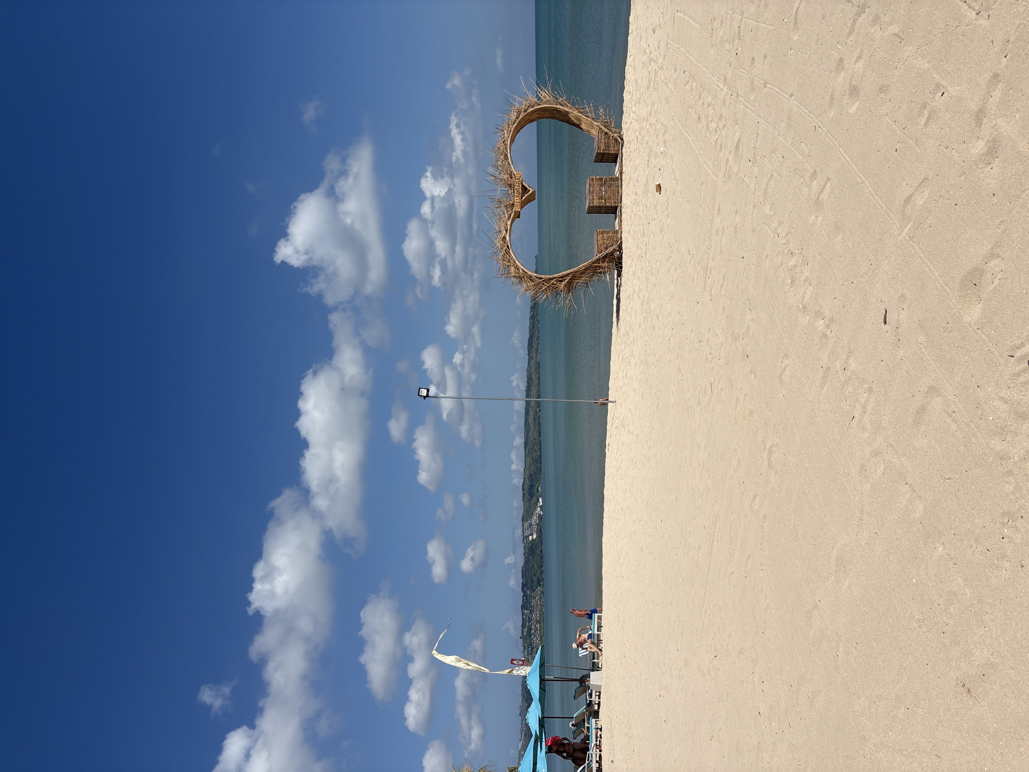 A bright, sunny day at Kuta Beach, Indonesia. The foreground is dominated by a wide expanse of light-colored sand with faint footprints and tire tracks. To the right, a large, heart-shaped structure made of natural materials, likely dried grass or reeds, sits on the sand. Inside the heart, two small, cube-shaped seats flank a low table. Beyond the sand, the calm, turquoise ocean stretches to the horizon. In the distance, a lush green coastline is visible, dotted with buildings and hills. The sky is a vibrant blue, punctuated by fluffy white clouds. On the left side of the image, several turquoise beach umbrellas are set up, providing shade for lounge chairs where a few people are relaxing. A tall, white flag with a curved design flies from a pole near the umbrellas. A single streetlamp stands on a tall metal pole in the middle ground. The overall scene depicts a peaceful and inviting beach setting.