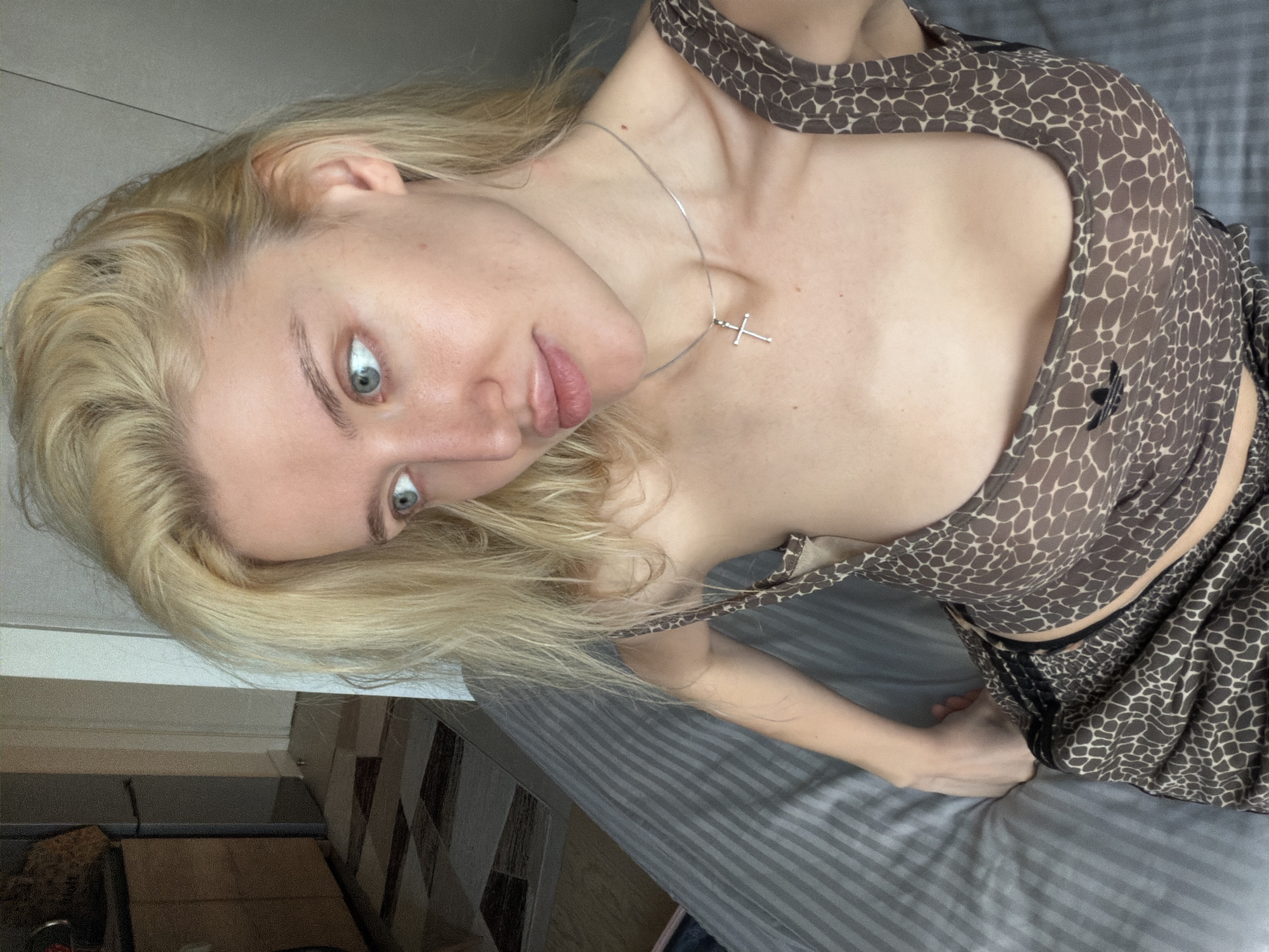 A blonde woman with pale skin and light eyes is looking at the camera. She is wearing a brown and white patterned top and a silver necklace with a cross pendant. She is indoors, possibly in a bedroom, as there is a bed with grey striped sheets visible in the background. There are also other interior design elements that might indicate the apartment is located in Belgrade. No city features are visible.