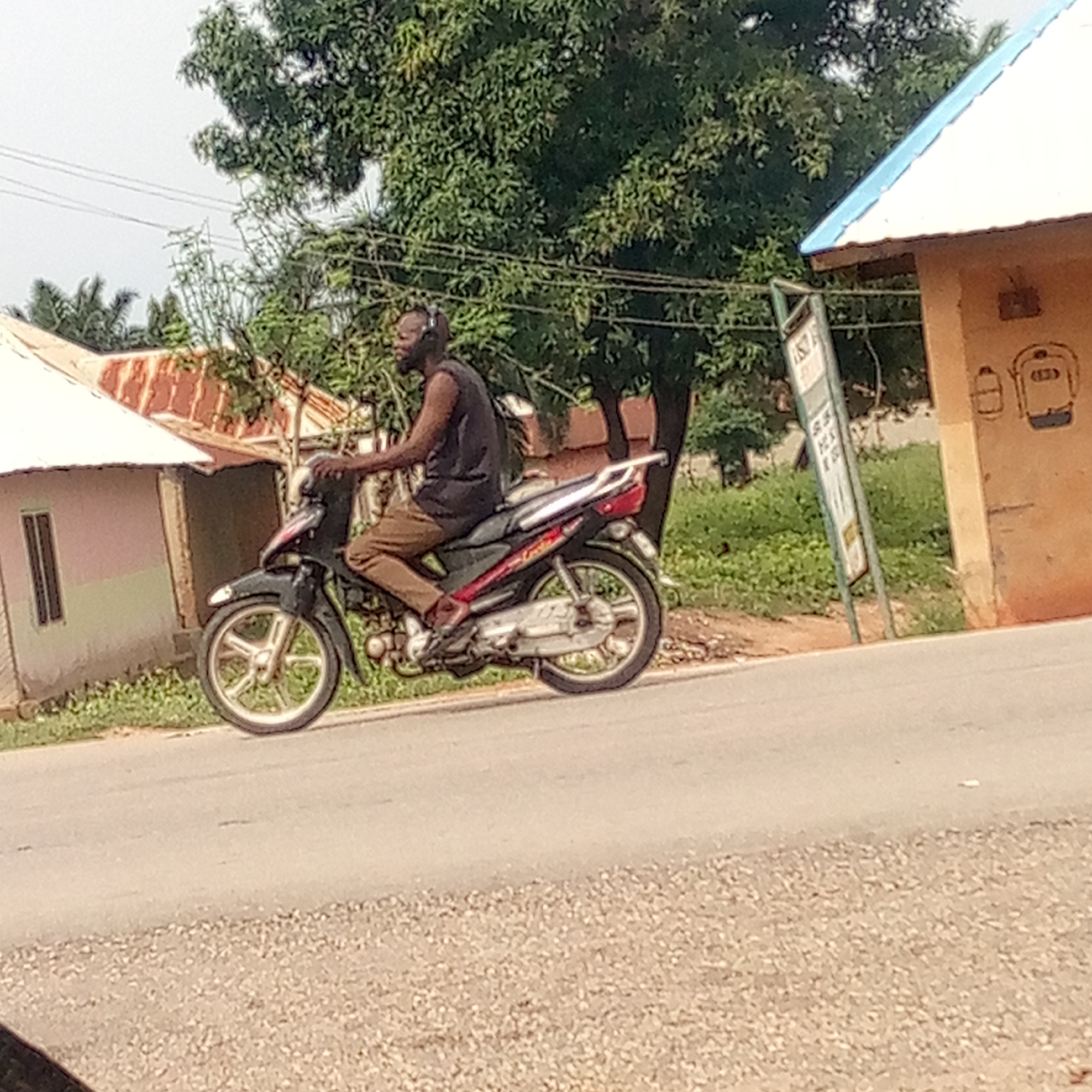 A candid outdoor shot captures a man riding a black and red underbone motorcycle on a paved road in Mararaba, Nigeria. The man, with dark skin, a beard, and short dark hair, wears a dark sleeveless top and light brown trousers. He appears to have an earpiece or headphone on his left ear and a slight smile. His feet are positioned on the footrests, and his hands are on the handlebars, indicating he is actively driving.
The road is asphalt or paved, bordered by a gravelly shoulder on the right. In the background, on the left, are two simple houses with corrugated iron roofs – one white and one rusty brown. Lush green vegetation and trees are abundant throughout the scene, suggesting a verdant environment.
On the right side of the road, a light brown building with a bright blue corrugated roof is visible. This building features simple white line drawings on its wall, one resembling a battery or container, and another a stylized bus or vehicle with illegible text (possibly "(ED)") inside. A tall, green signpost stands in front of this building; its text and graphics are mostly obscured and blurry, but white lettering is faintly visible. The general setting suggests a daytime scene, possibly with an overcast sky, as the lighting is soft without harsh shadows.