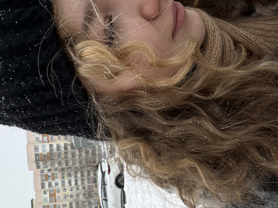 The image displays an outdoor scene during snowfall in the city of Nizhnekamsk, Russia. The primary subject is an individual, likely female, positioned in the right foreground, depicted from the left side of their face and upper body. Her light brown to blonde curly hair and black knitted hat are visibly speckled with small white snowflakes. She wears a light brown knitted scarf wrapped around her neck. Her right eye is partially obscured by hair, and her gaze appears directed downwards or slightly to the right. The bridge of her nose and part of her mouth (lips) are visible.
The background reveals a snow-covered urban landscape. White snow blankets the ground, and several vehicles are parked, partially obscured by the snow. A prominent white SUV is visible in the mid-ground on the left, facing left. In the far background, two multi-story residential apartment buildings, light in color (beige/pale pink tones), stand under an overcast, light grey sky. Bare deciduous trees are interspersed within the scene.