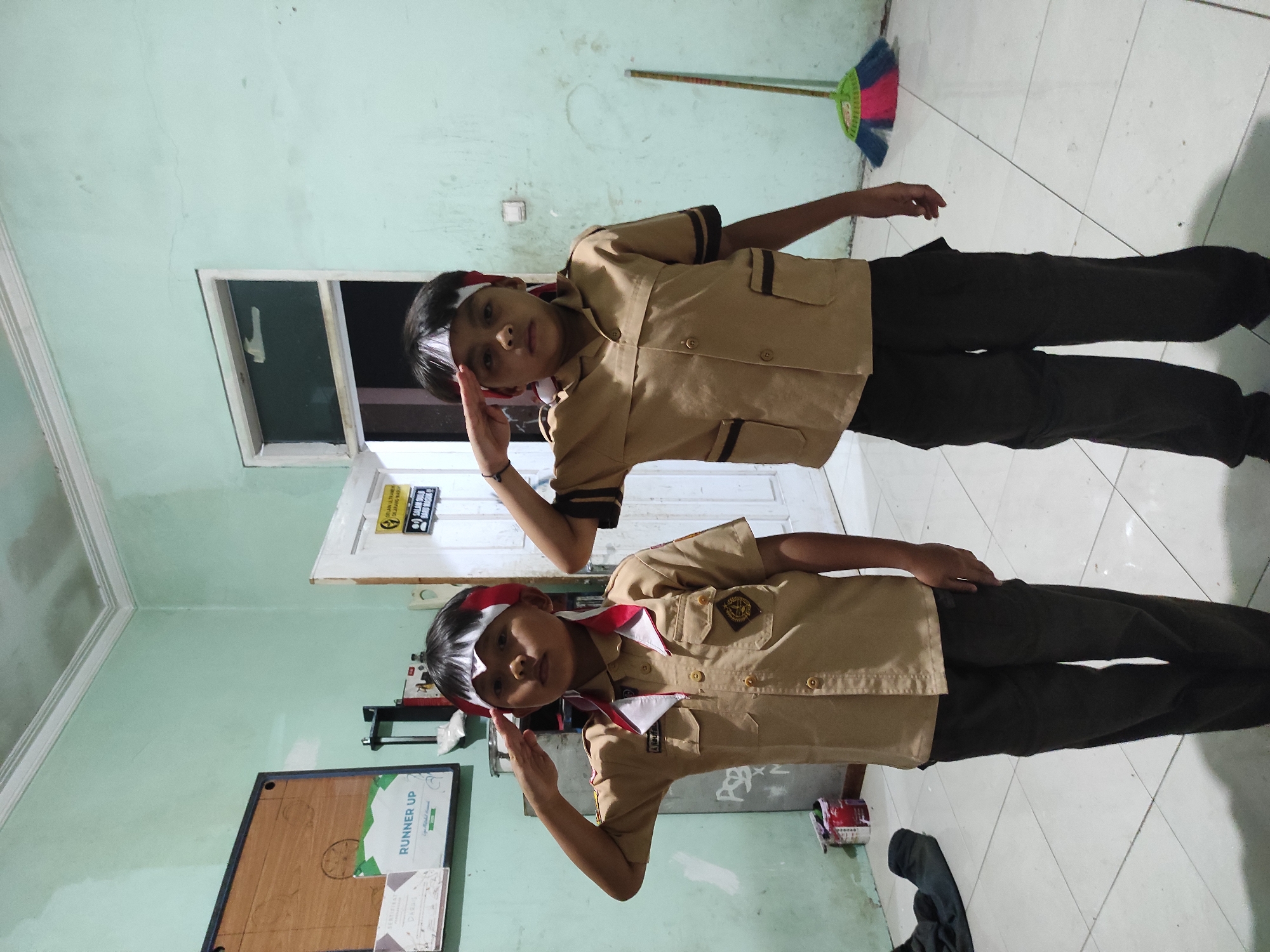 Two young boys, likely elementary school age, stand facing forward in what appears to be an indoor space. They are both wearing khaki-colored uniforms with red and white trim around the collars and cuffs. Both boys have a white band with red accents tied around their foreheads and are performing a salute, with their right hands raised to their foreheads. The boy on the left is slightly closer to the camera and has a small emblem on his left breast pocket. The boy on the right is positioned behind and to the right of the first boy.
The environment is a room with light green walls and a tiled floor. To the left, a bulletin board is mounted on the wall, displaying what looks like certificates and a sign that says "RUNNER UP". There is a doorway behind the boys, partially open, revealing a dark interior. To the right, a broom with colorful bristles (pink, green, and blue) leans against the wall. Various items are visible in the background, including what appears to be paint cans and other miscellaneous objects. The overall lighting suggests an indoor setting, possibly a school or community building.
The location is the city of Sumedang, Indonesia.