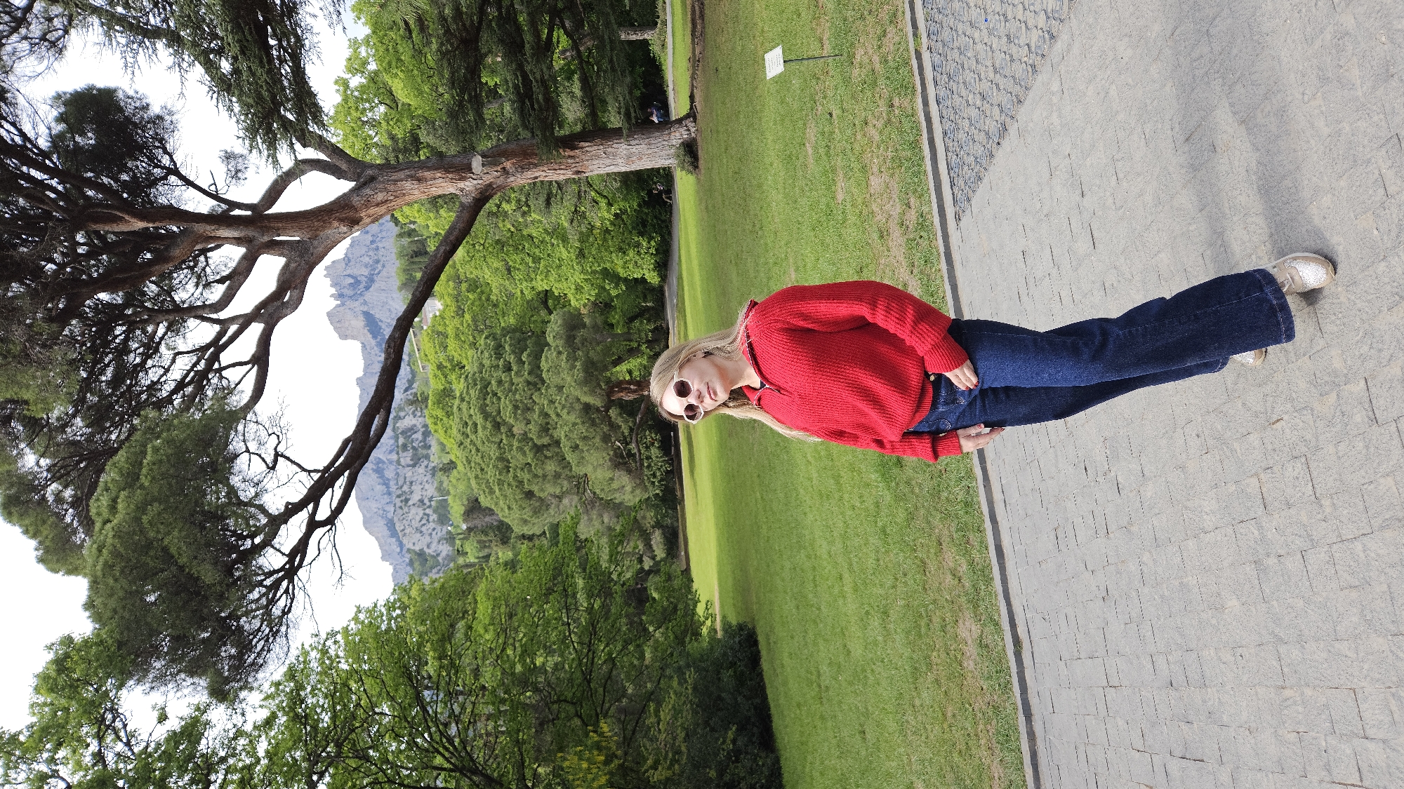 A young, fair-skinned woman stands on a paved walkway next to a large grassy area. She wears a red sweater, blue jeans, and light-colored shoes, and also sports sunglasses. Behind her is a landscape featuring a large tree with dark green foliage to the left and other trees and green vegetation extending towards a rocky mountain formation in the background. The sky appears overcast or bright with muted lighting. The setting is located in Unknown, Unknown.