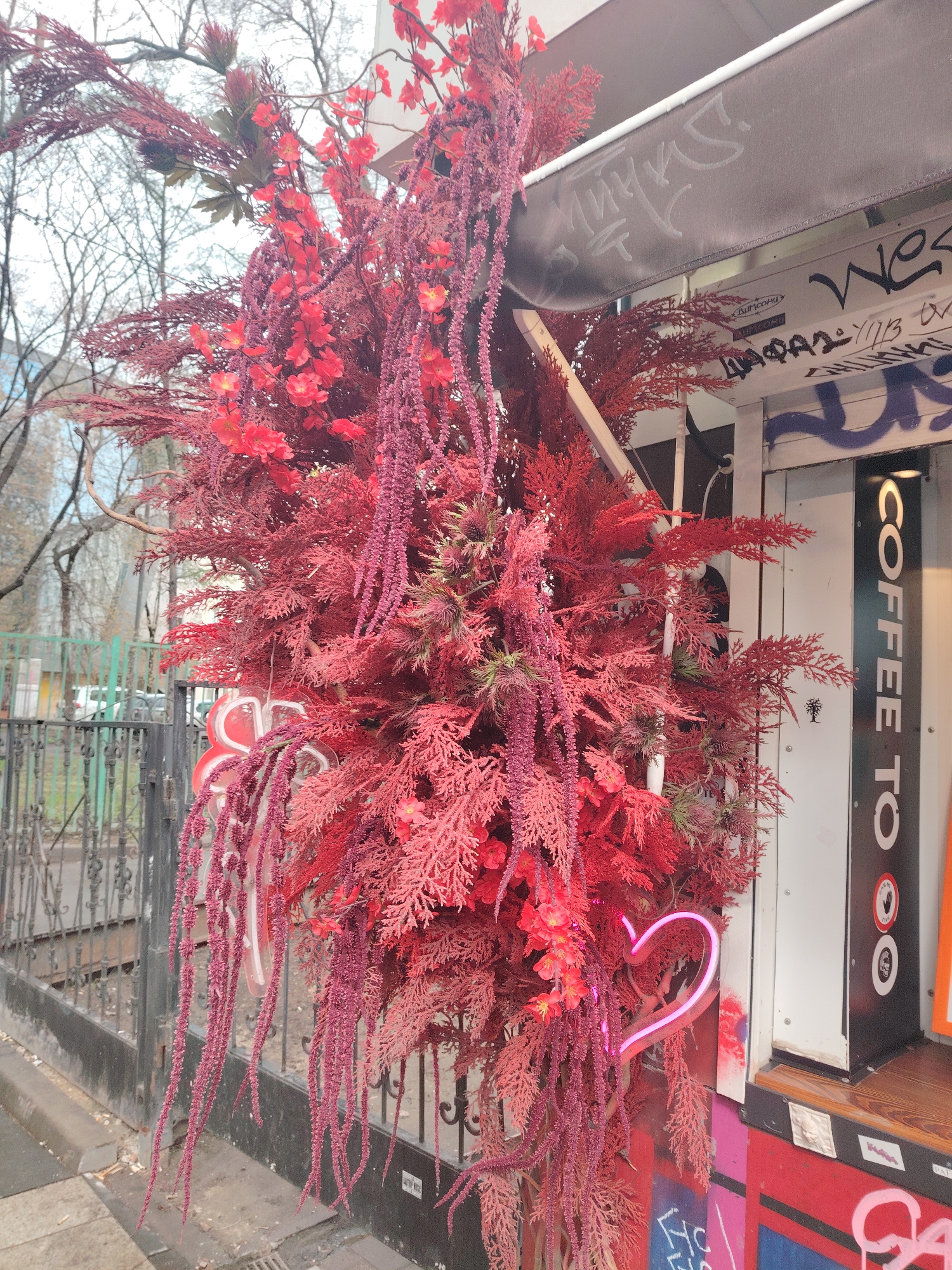 The image captures a vibrant and slightly surreal street scene, likely in a European city. A dense arrangement of red foliage, including cascading amaranthus caudatus, obscures part of a building facade. This building features graffiti, a striped awning, and a sign that reads "COFFEE TO". A neon heart-shaped light is placed within the foliage. The image appears to be from Moscow.