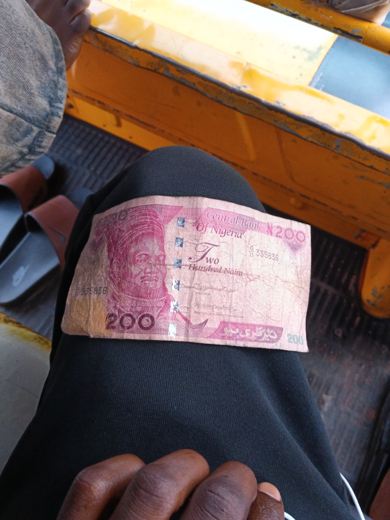 A Nigerian 200 naira note is held by a person's hand. The banknote is pink and features a portrait of a man. The note is partially crumpled and worn. The person holding the note is wearing dark clothing. In the background, a yellow vehicle with a weathered texture is visible, along with a dark, textured surface that appears to be the floor or seating area of the vehicle. Two brown sandals are also visible in the background, to the left of the banknote. The lighting suggests an outdoor or dimly lit indoor setting. The location is Unknown, Unknown.