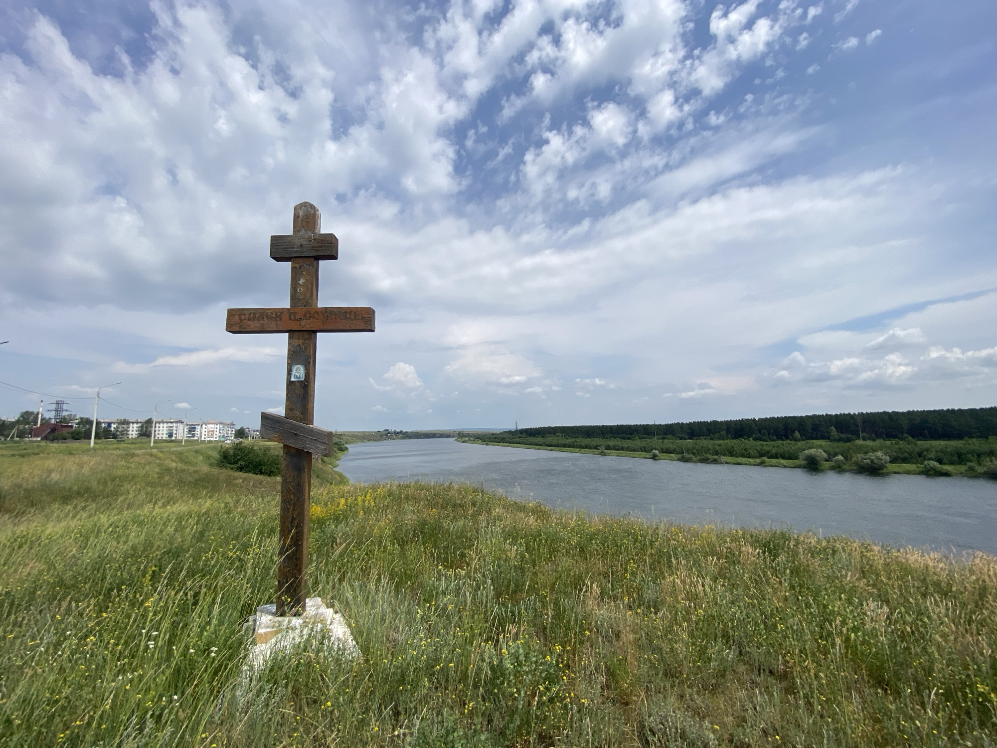 An outdoor, eye-level, medium shot captures a wooden cross standing on a grassy hill overlooking a wide river. The cross is made of weathered dark wood, with a plaque or inscription visible on its vertical beam. The hill is covered in tall, green grass with scattered yellow and white wildflowers. The river, a muted blue-grey, flows from left to right across the frame, with a dense, dark green forest lining its far bank. The sky is a bright blue with scattered white, fluffy clouds. In the distance, on the left side of the image, a cluster of multi-story buildings and power lines suggests the presence of a settlement. The scene is tranquil and evokes a sense of landscape and natural beauty. The image does not contain any people.