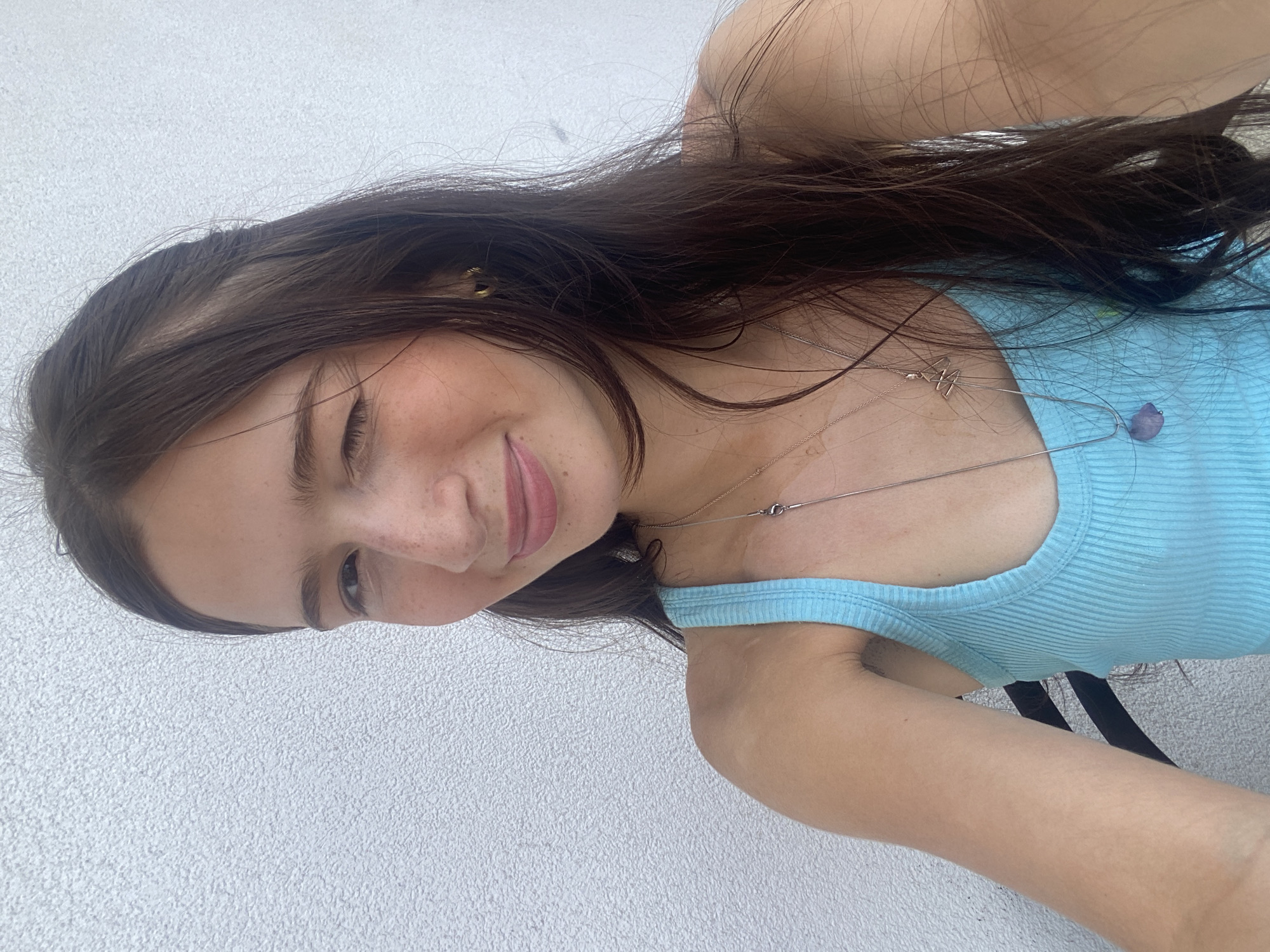 A young woman is positioned at an angle against a pale textured background, likely an exterior wall. She is looking downward, with a slight smile. She wears a light blue ribbed tank top and two delicate silver necklaces. Her dark, long hair falls loosely around her shoulders. The environment appears to be outdoors, suggested by the natural lighting and the wall texture. No information regarding the city of London or the United Kingdom is visibly present in the image.