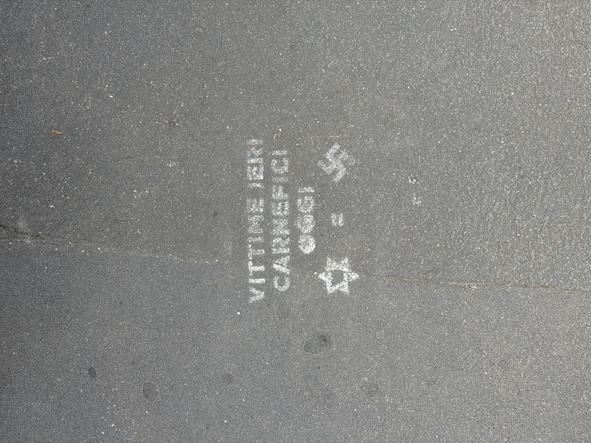 The image depicts street art on asphalt. The text "Vittime ebrei caruccici" is vertically stenciled, and next to it is the Star of David and the Swastika. The location is Milan, Italy.