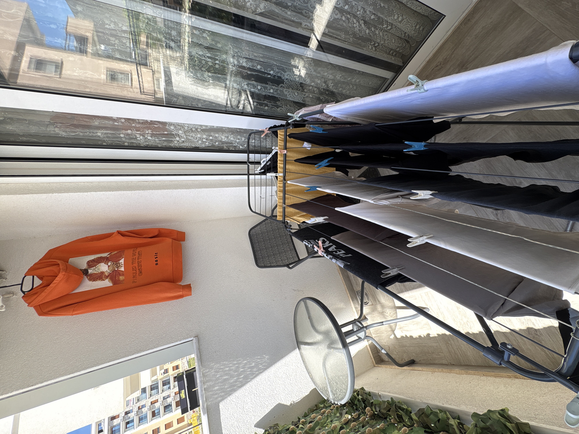 The image depicts a balcony setting. A clothes drying rack is laden with laundry, secured by clothespins. Clothes of varying colors, including white, gray, and black, hang from the rack. An orange hoodie, printed with a graphic design, hangs on the wall, supported by a coat hanger. A metal chair and glass-topped table are positioned on the balcony floor. In the lower portion of the frame, vegetation is visible, likely potted plants. Through a window, a building can be seen. Based on the architectural style visible in the window reflection, it is deduced that the scene is located in the city of Bar, Montenegro. No people are visible. The scene appears to be a residential setting where laundry is being air-dried on a balcony.