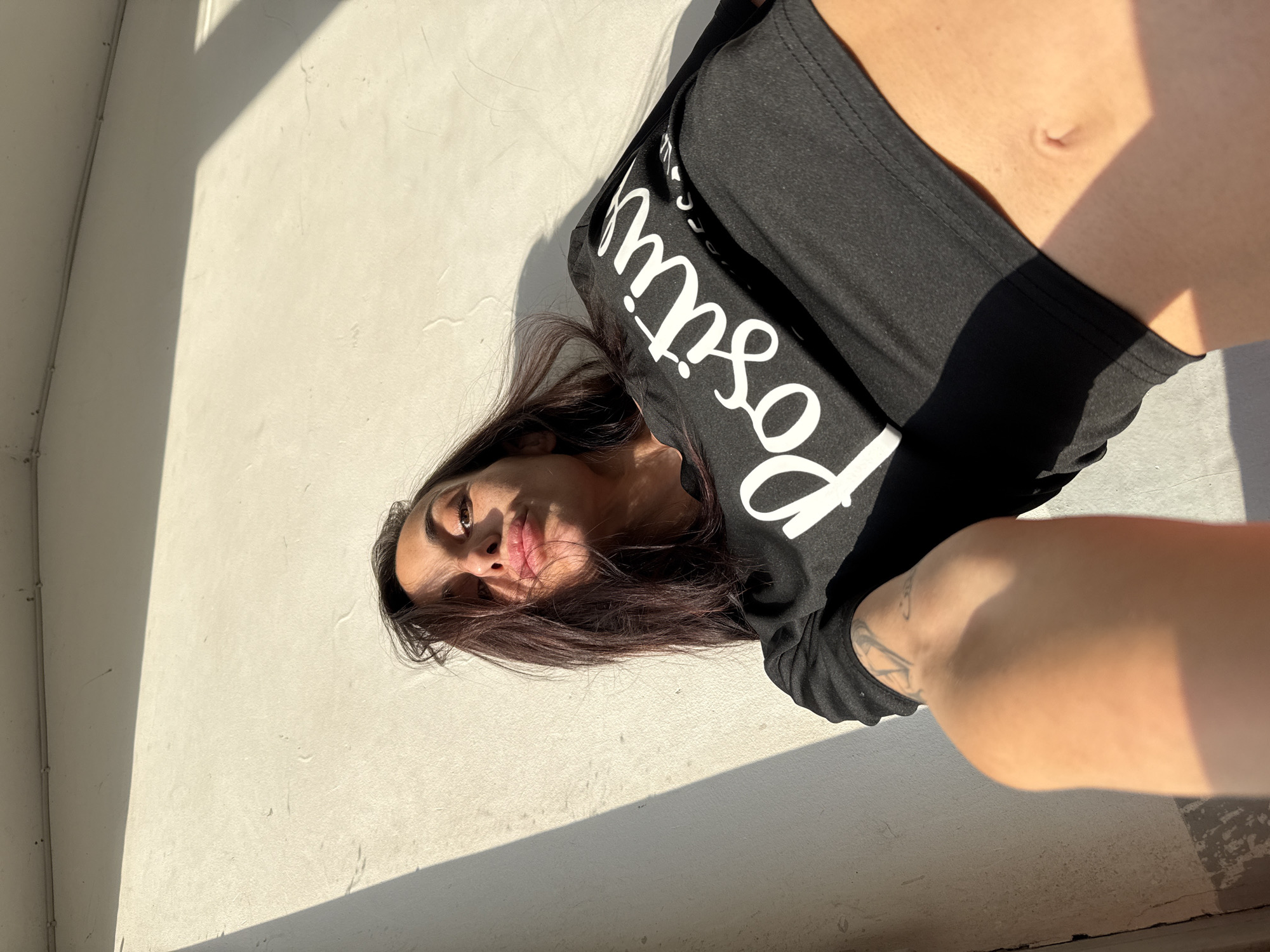 A woman is lying on a bright, sunlit surface. She has dark hair and is wearing a black shirt with the word "Positive" printed on it. Part of her torso is visible, along with her arm which has a tattoo. The environment appears to be a wall next to her body, with a beam of sunlight casting a shadow. The specific location in Pleven, Bulgaria, cannot be determined from the image.