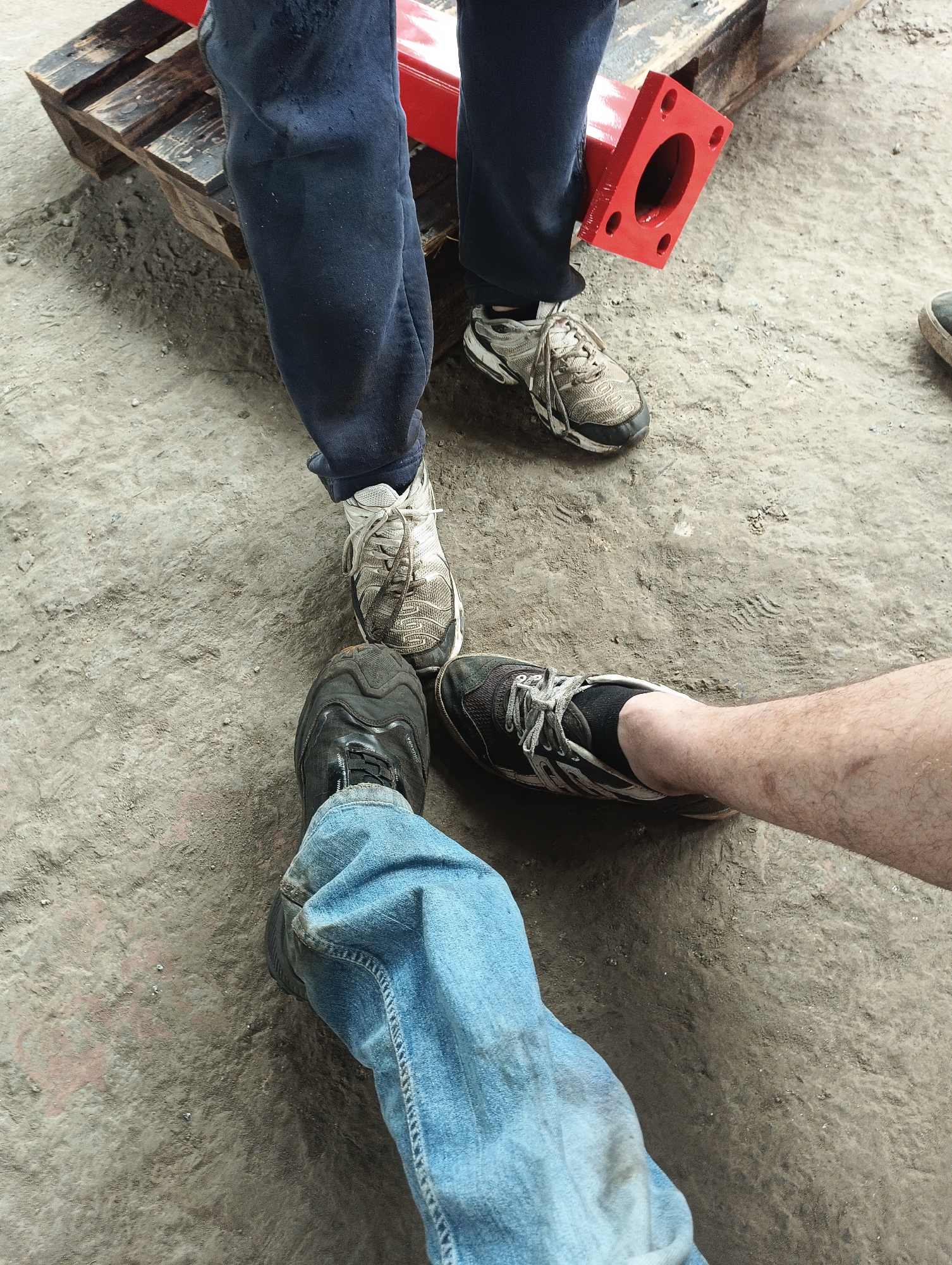 The image presents a ground-level view of three individuals standing on a concrete surface. The individuals are identifiable only by their lower legs and footwear. One person is wearing blue jeans and black work boots. Another is wearing navy blue sweatpants and gray and white running shoes. The third individual is wearing shorts and black socks with gray and white running shoes. A wooden pallet with red metal component rests behind the person in navy blue sweatpants. The setting appears to be an indoor industrial or garage area.