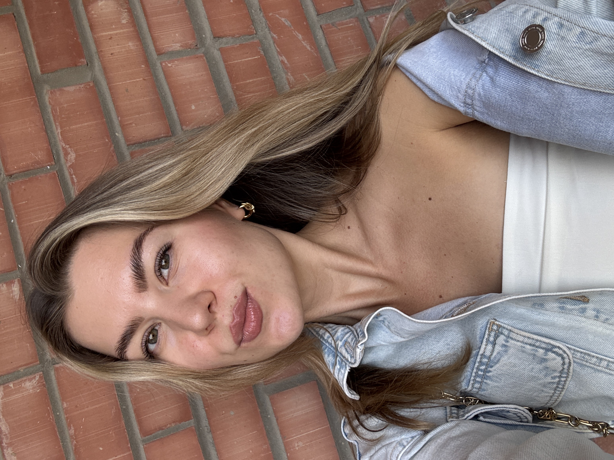 A young Caucasian woman with long, blonde hair is taking a selfie outdoors, with a red brick wall as the background. She is wearing a light blue denim jacket, unbuttoned, revealing a white strapless top. She has a gold hoop earring in her left ear. Her face is turned slightly towards the camera, and she is puckering her lips. She has light green eyes and well-groomed eyebrows. The lighting appears to be natural, suggesting daylight.