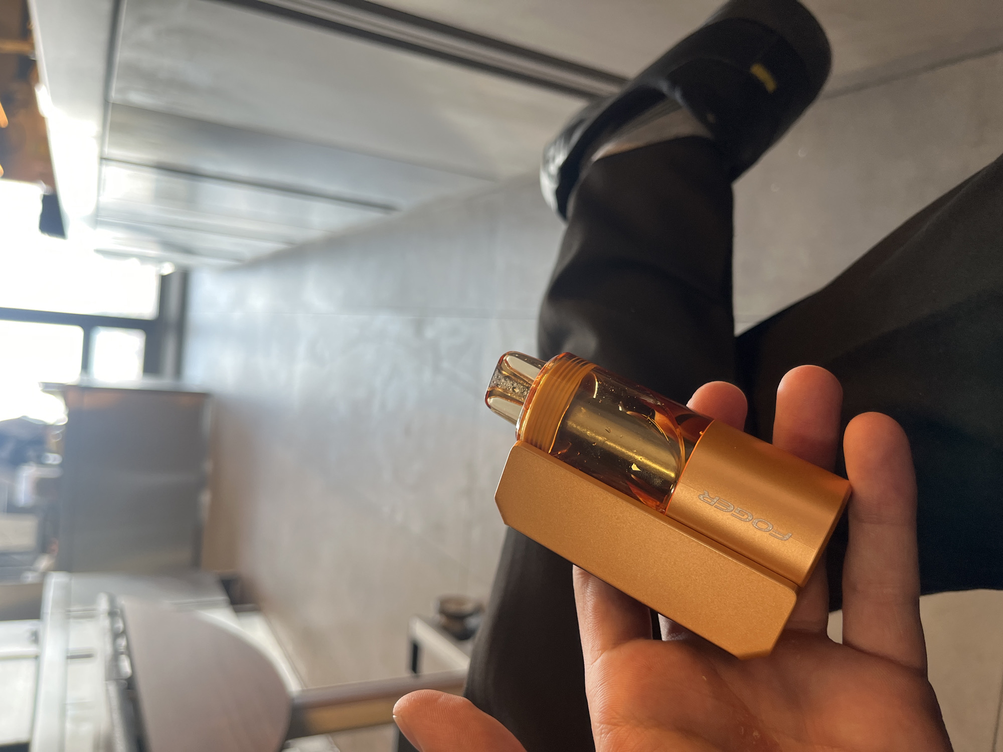 The image, taken in Sofia, Bulgaria, displays a person's hand holding a golden-orange vape device in an indoor setting. The device is compact and rectangular, featuring a clear tank filled with an amber-colored liquid. The brand name "FOGGER" is prominently visible in white lettering on the side of the device. The hand holding the vape is fair-skinned.
In the soft-focus background, parts of a person wearing black trousers and a black shoe are visible on the right, suggesting the individual holding the device is seated. The setting appears to be a modern interior, possibly a cafe or office, brightly illuminated by natural light entering from a large window on the left side of the frame. Blurred architectural elements include light-colored walls, ceiling panels, and indistinct furniture, indicating a contemporary environment during daylight hours. No specific activities beyond holding the device are depicted, and no emotions are discernible.