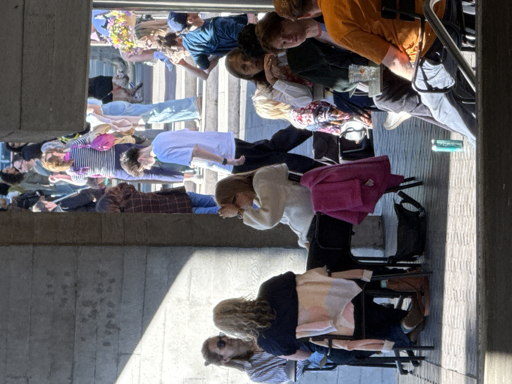 The image, taken in London, United Kingdom, depicts a lively outdoor scene with numerous individuals, primarily young adults, gathered at tables, likely an al fresco dining or social area. The perspective looks down onto the group from a slight elevation, with the scene illuminated by strong sunlight causing distinct shadows.
In the foreground, two individuals are seated, one with curly blonde hair and sunglasses pushed onto their head, wearing a dark top, and another in a striped shirt. Further back, a person in a beige top and black pants is visible, alongside another with a vibrant pink backpack beside them. The tables extend into the midground, occupied by more people. A prominent figure in the upper-middle section wears a white shirt and black trousers, while another has a decorative floral wreath on their head. A small, light-colored dog is visible beneath a table towards the upper left, partially hidden.
People appear to be socializing, eating, and drinking. Various objects are scattered on and around the tables, including clear glasses, a green can, and handbags. The ground is paved with a textured, brick-like pattern, and sturdy concrete walls or pillars frame the scene, suggesting an underpass or architectural overhang. The bright sunlight and long shadows indicate a clear, sunny day, likely morning or afternoon. Most individuals appear relaxed and engaged in conversation. The text on the green can is too small and blurred to be legible.