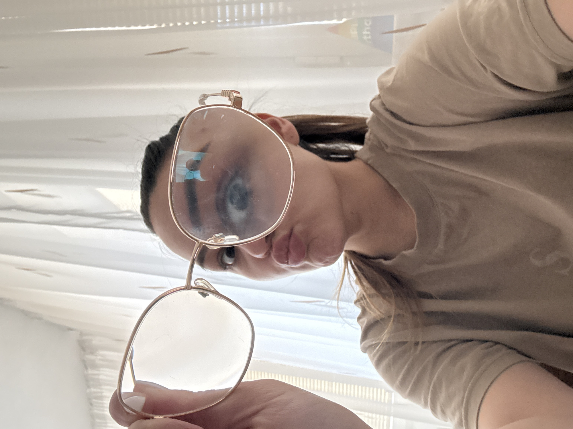 A woman with dark hair is holding a pair of gold-rimmed glasses in front of her face. She is wearing a beige t-shirt. The background is blurred but appears to be a room with white window blinds. There is insufficient information to determine if this image was taken in the city of Sofia, Bulgaria.