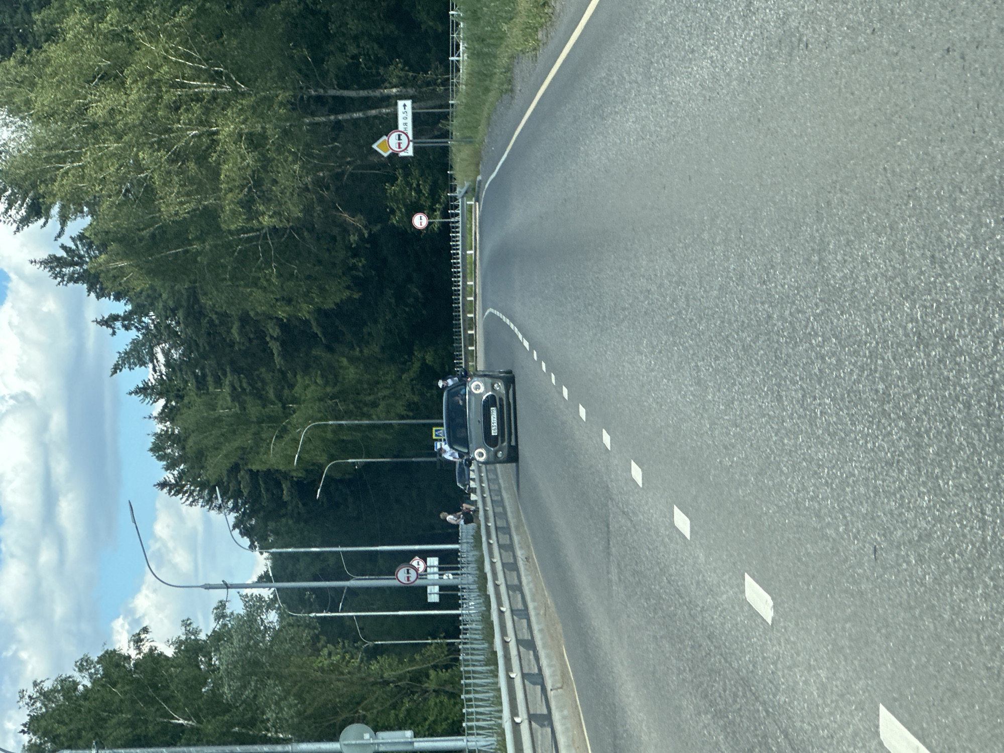 The image shows a gray Mini Cooper driving on a paved road with dashed white lane markings. The car is in the center of the frame, facing forward. The road appears to be a highway or a major road, bordered by a metal guardrail on the left and trees on both sides. The sky is cloudy, with patches of blue. Several streetlights are visible along the left side of the road. There are traffic signs visible on the right side of the road, including a diamond-shaped sign with two cars and an arrow, and a circular sign with a red border and a horizontal bar. The location is identified as Lukovnya, Russia. The overall scene depicts a sunny day on a road surrounded by nature.
