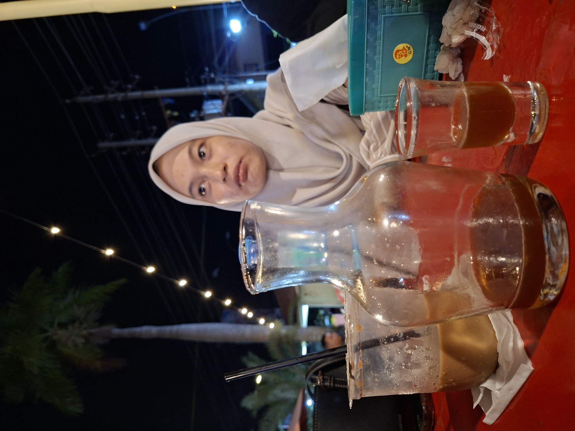 At night in Lhokseumawe, Indonesia, a woman wearing a light gray hijab sits at a red table. On the table are a glass of dark liquid, a pitcher with some liquid remaining, a plastic cup with a straw, and a blue plastic container. String lights illuminate the area above a palm tree. In the background are utility poles and distant lights.