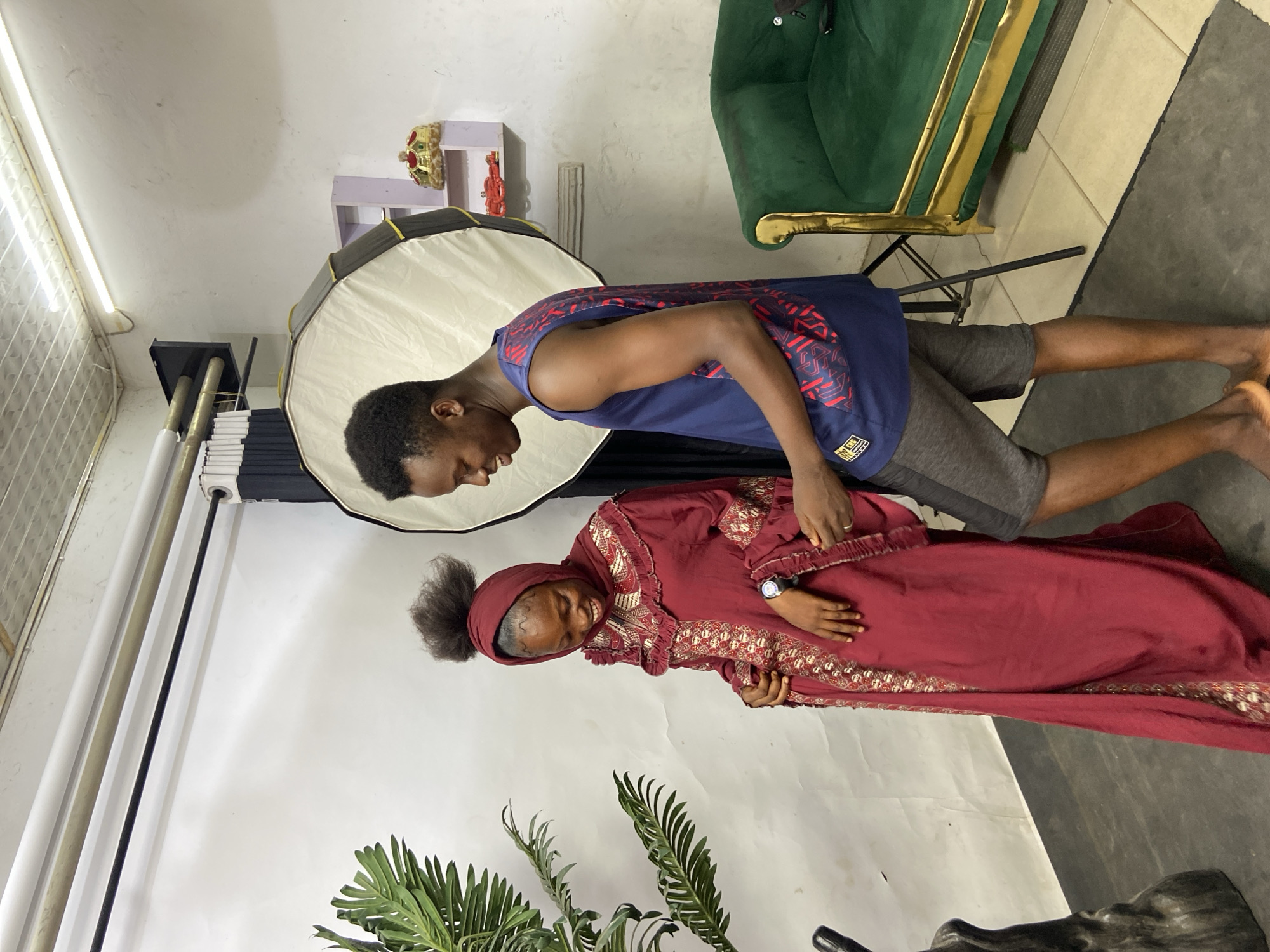 In a studio setting in Mararaba, Nigeria, a man and a woman pose for a photograph. The man, wearing a blue sleeveless shirt and grey shorts, stands near the woman, who is clad in a maroon dress and head covering. A large, white umbrella light diffuser is positioned to the side of the subjects. A green chair with gold accents, a potted plant, and a shelf with decorative items can be seen in the background.