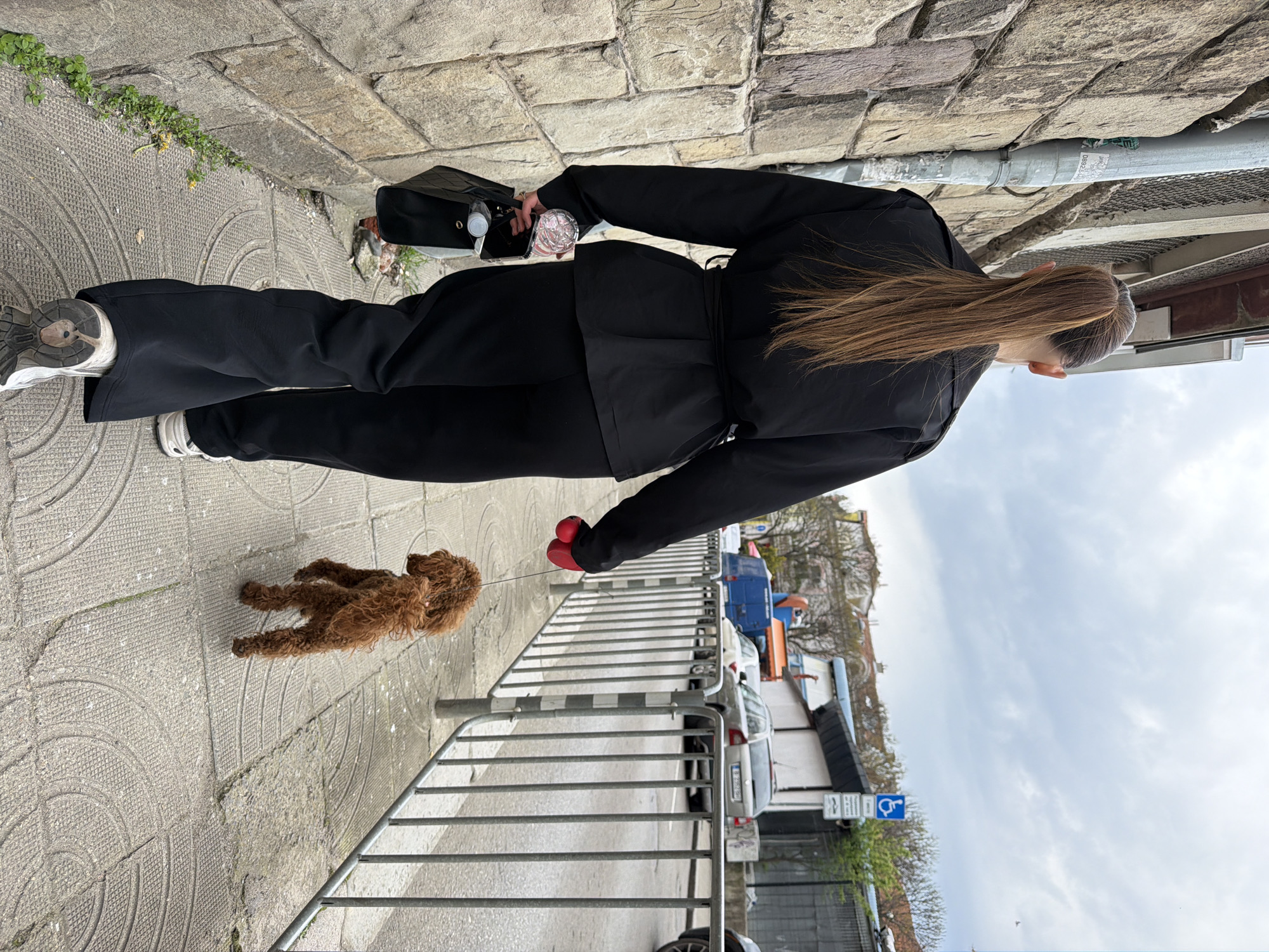 The image, rotated counter-clockwise, depicts a person walking a small dog on a leash along a paved sidewalk in Sofia, Bulgaria. The person, viewed from behind, is dressed in an all-black ensemble: wide-leg trousers and a belted jacket. Their long, light brown hair is tied back, and they wear light-colored sneakers with thick soles. They carry a black handbag in their left hand, which appears to hold a white bottle and a clear cup. A vibrant red leash extends from their right hand to a fluffy, reddish-brown dog, resembling a poodle, walking ahead on the sidewalk.
The concrete sidewalk features circular patterns and scattered patches of green weeds growing in its cracks. To the person's left, a rustic stone wall is constructed from large, irregular blocks, with a grey vertical pipe running along its length; indistinct text is visible on the pipe. To their right, a metal barrier separates the path from a street with parked vehicles, including a silver car and a partially visible blue van. Buildings with light-colored facades and dark roofs line the street beyond. The sky above is overcast, suggesting a daytime scene. A blue sign displaying a handicapped parking symbol and other unreadable white text is visible near the street.