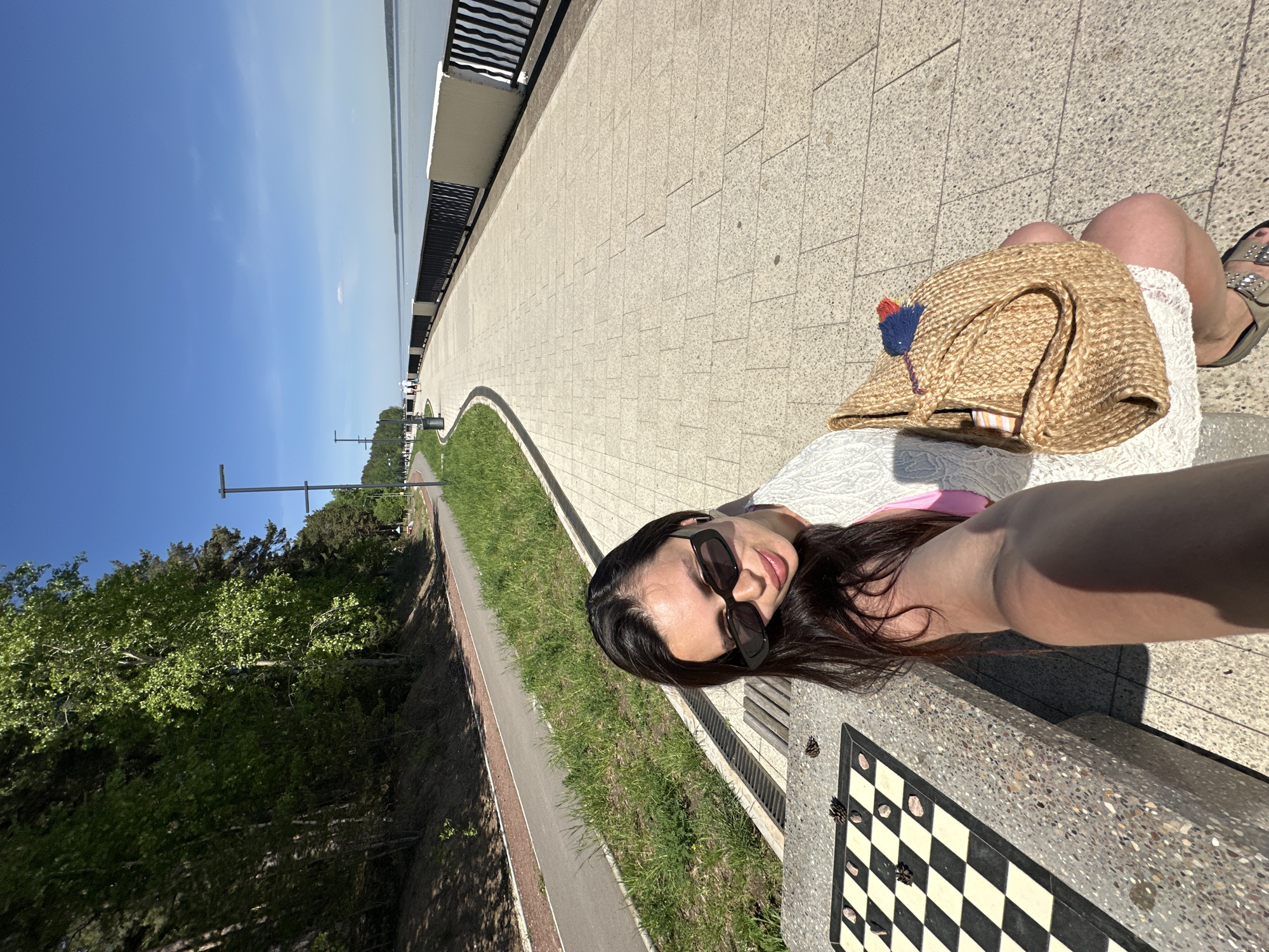 A young woman with long, dark hair, wearing sunglasses, a white dress, and sandals, is taking a selfie while sitting on a concrete bench with a built-in chessboard. A straw bag with blue and red tassels rests next to her on the bench. The bench is located on a paved pathway along a waterfront in Chaikovsky, Russia. To the left of the pathway is a grassy area with trees, and to the right is a higher paved area with a metal railing overlooking the water. The sky is blue with some light clouds. Several pinecones are scattered on the chessboard.