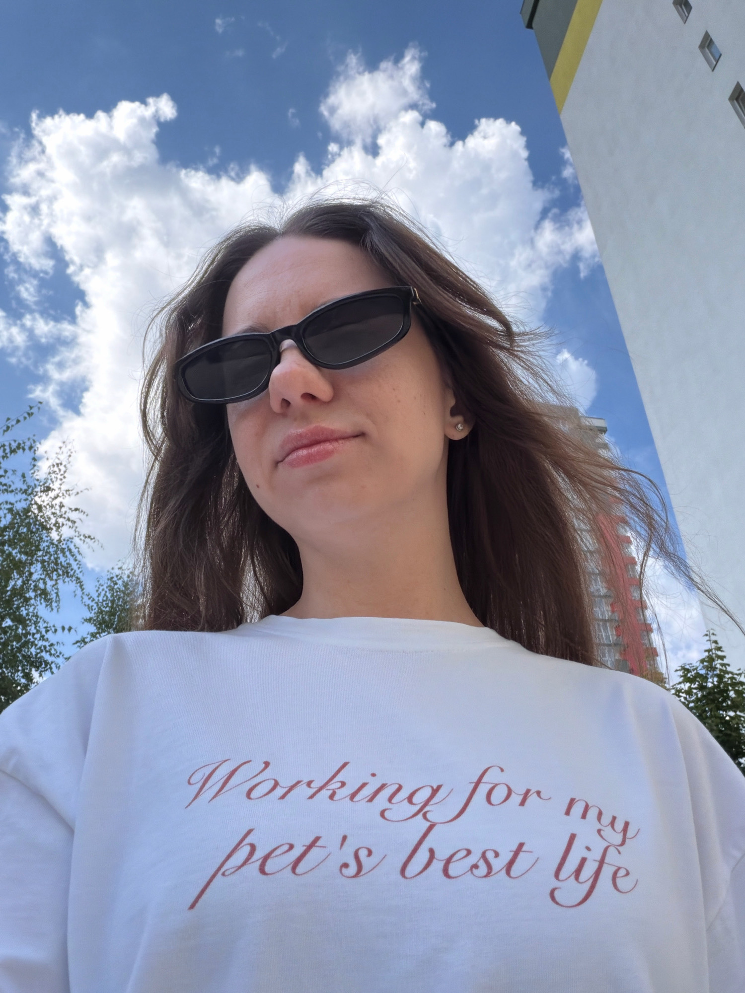 A young woman wearing sunglasses and a white t-shirt with the text "Working for my pet's best life" in red script stands outdoors, looking up at the sky. Her brown hair is blowing in the wind. In the background, a modern white and yellow building is visible, with a hint of a reddish-orange facade on another building to the right. The sky is blue with fluffy white clouds. Trees are also visible on the left and right edges of the frame. The scene is likely in Kyiv, Ukraine, given the architectural style and the mention of a city location.