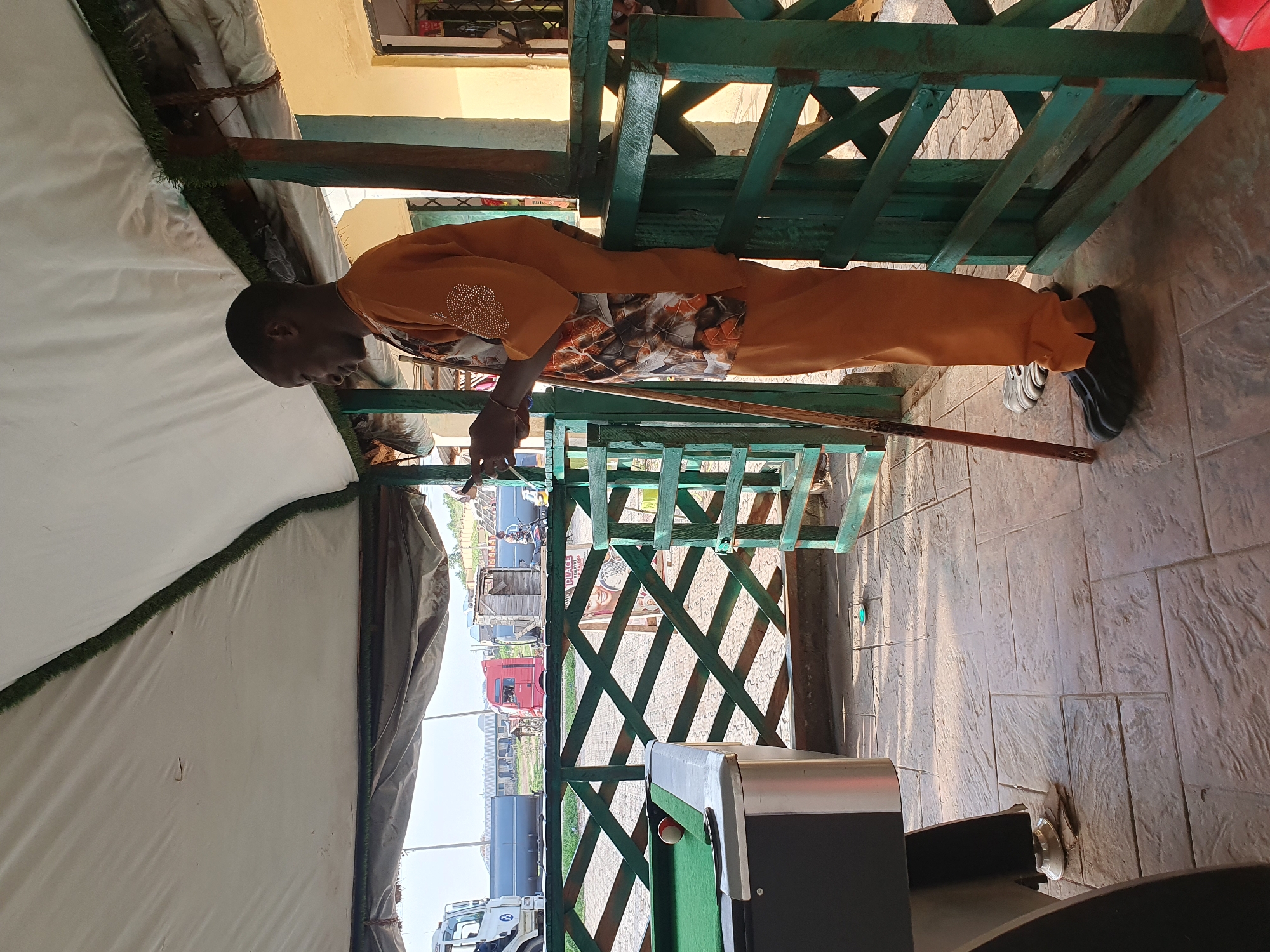The image depicts an outdoor recreational area, possibly a bar or a place for games, situated in Lokoja, Nigeria. A man is standing by a green wooden railing, looking at his smartphone. He is wearing an orange outfit with a patterned shirt. A pool table with a green felt surface and one ball is visible to the left. In the background, there are trucks and buildings, suggesting an urban or semi-urban setting. The area is covered by a white canopy, providing shade. The ground is paved with textured concrete tiles.