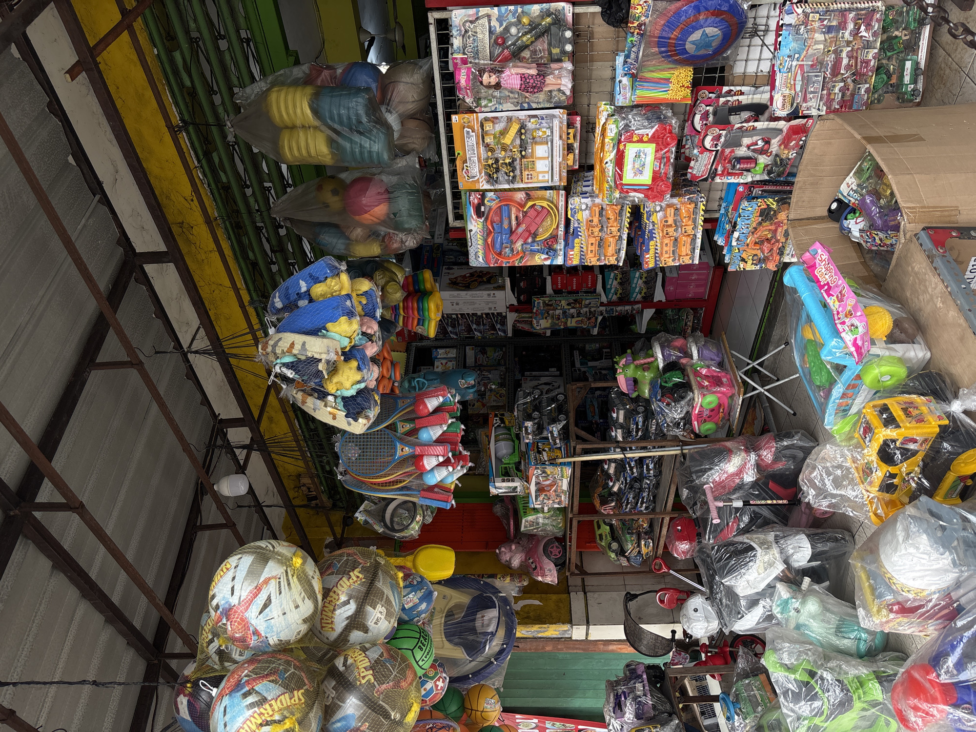 This is a full shot of a street-side toy shop in Denpasar, Indonesia. The shop is cluttered with a variety of toys displayed both inside and outside. In the foreground, several large toys are covered in plastic packaging, including a yellow toy truck, a red and blue toy car, and a green plastic ride-on toy. To the left, smaller plastic-wrapped toys such as a purple car and a green scooter are visible. In the middle ground, several toy motorcycles are also plastic-wrapped. Behind the motorcycles, shelves are stocked with more boxed toys, including toy construction vehicles, toy trucks, and toy airplanes. A doll is also visible on a shelf. Hanging from the ceiling are numerous colorful balls, many of which appear to be sports balls with cartoon character designs such as Spiderman. Other items hanging include rackets and shuttlecocks, as well as bags of stacked colorful plastic rings. The shop appears to be under a metal awning with yellow and green corrugated metal siding. The overall scene depicts a busy and vibrant toy store, likely catering to children.